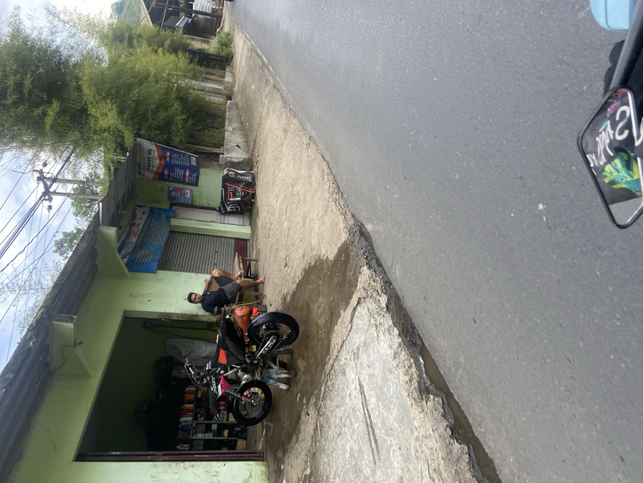 The image shows a street in Cipatat, Indonesia (latitude: -6.799, longitude: 107.444). A man in a black shirt and pants is sitting on a wooden crate in front of a building. A black and orange motorcycle is parked near him, with a green and black motorcycle in the foreground of the image. The building has a green facade with a closed metal door. There are some signs on the building in Indonesian. The street is paved and appears wet. There are a few small puddles in the road. The road is sloping slightly downhill to the right. There are some green plants in the background. The day is cloudy and overcast.