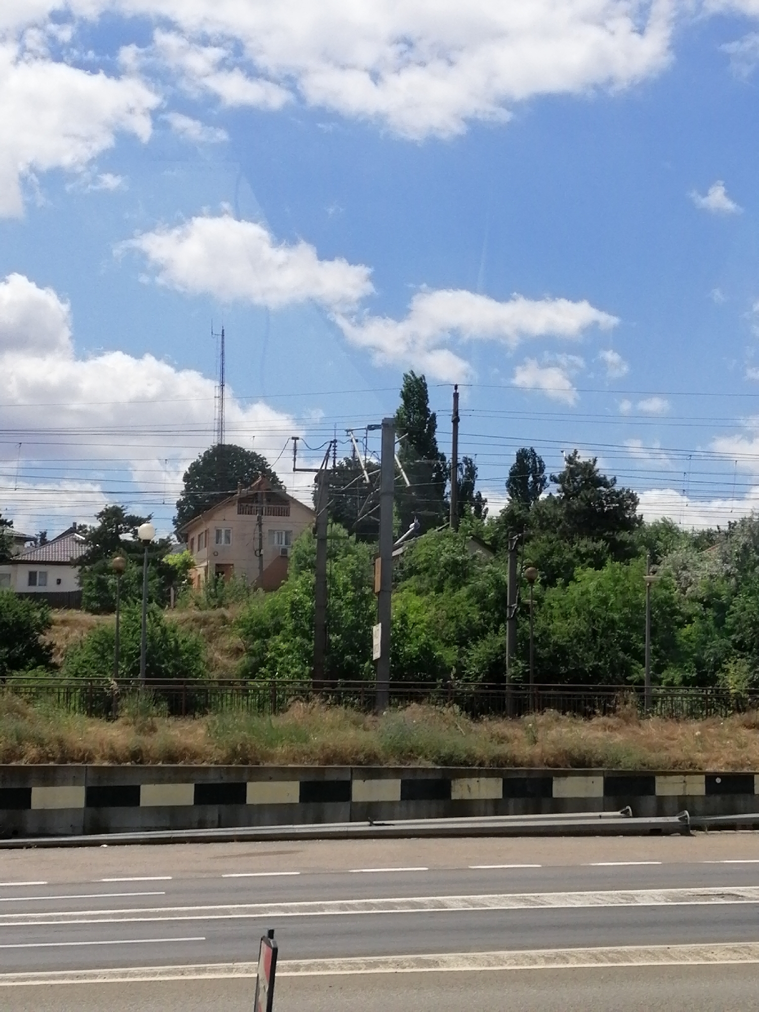 The image depicts a view of Fetești, Romania, featuring a residential area with houses nestled amidst lush greenery and trees. Overhead power lines crisscross the sky, indicating the presence of a railway or an electrical grid. A tall transmission tower rises above the houses, hinting at communication infrastructure. The foreground shows a road with white lane markings, suggesting a transportation route. The scene is bathed in daylight under a partly cloudy sky, evoking a tranquil, suburban atmosphere.
