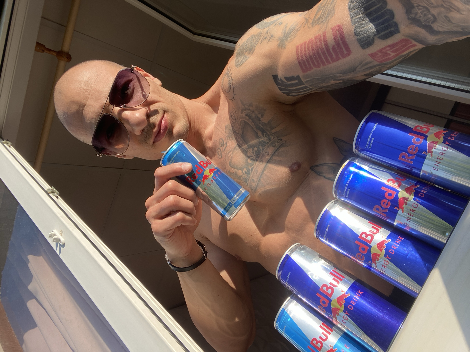 A shirtless, bald man wearing sunglasses is holding a can of Red Bull in his left hand. Several other cans of Red Bull are lined up in a row on what appears to be a white windowsill. The man has tattoos on his chest and both arms. The image is taken from a low angle, looking up at the man. The location appears to be inside a building in Pleven, Bulgaria, based on the Cyrillic text on the cans.