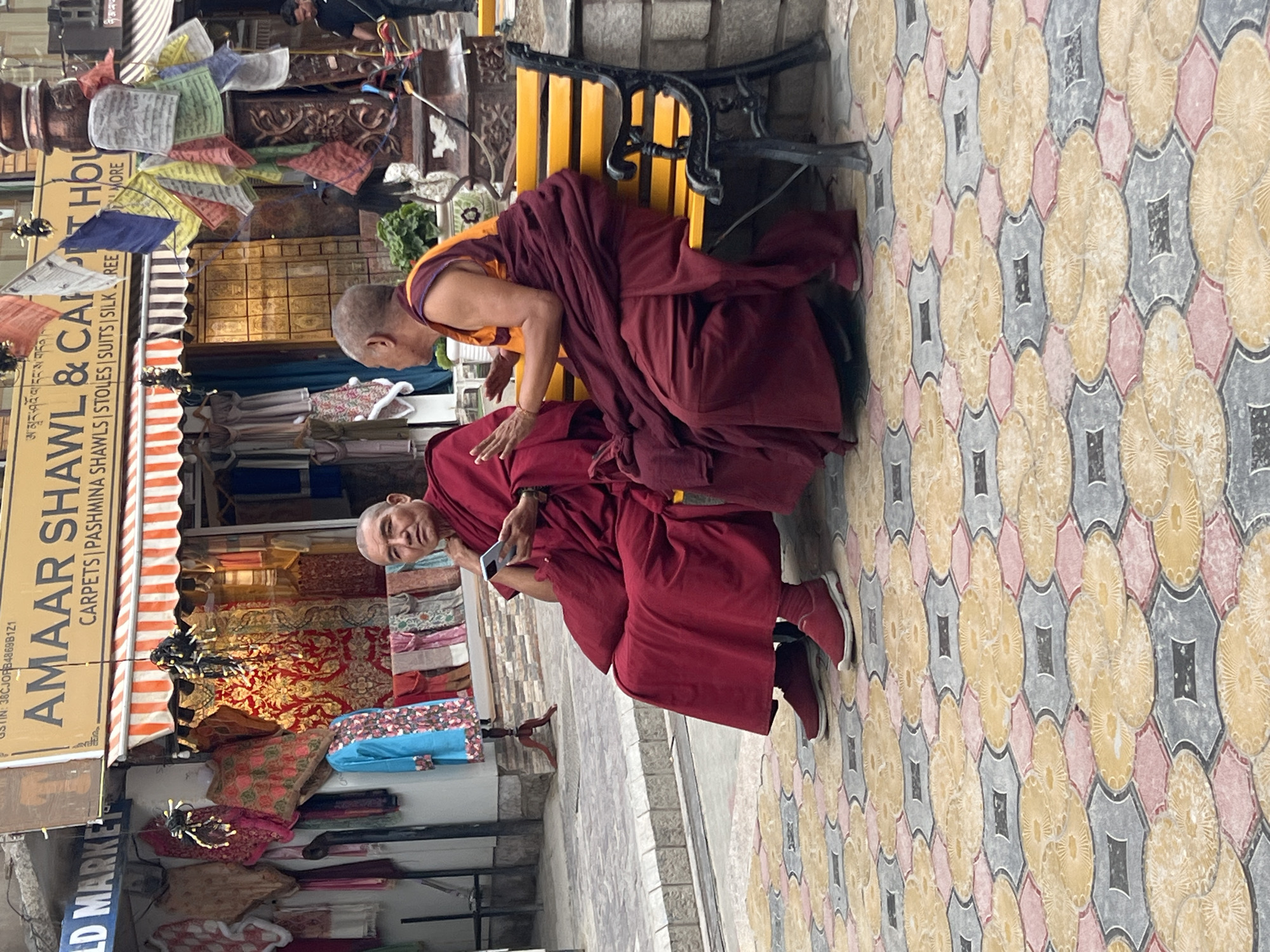 Two Buddhist monks are sitting on a yellow bench in Leh, India. The monk in the foreground is wearing a maroon robe and holding a smartphone. The monk behind him is wearing a similar robe and is leaning forward, looking down. The background shows a shop with prayer flags hanging outside and a display of colorful textiles. The ground is paved with a patterned tile. The scene appears to be taking place during the daytime. The monks seem relaxed, and the overall atmosphere is peaceful. Visible text includes "AMAAR SHAWL & CAR HOL" and "CARPETS | PASHMINA SHAWLS | STOLES | SUITS | SILK REE", which indicates the name and type of goods sold at the shop. The setting is a street in Leh, a popular tourist destination known for its Buddhist monasteries and vibrant culture.
