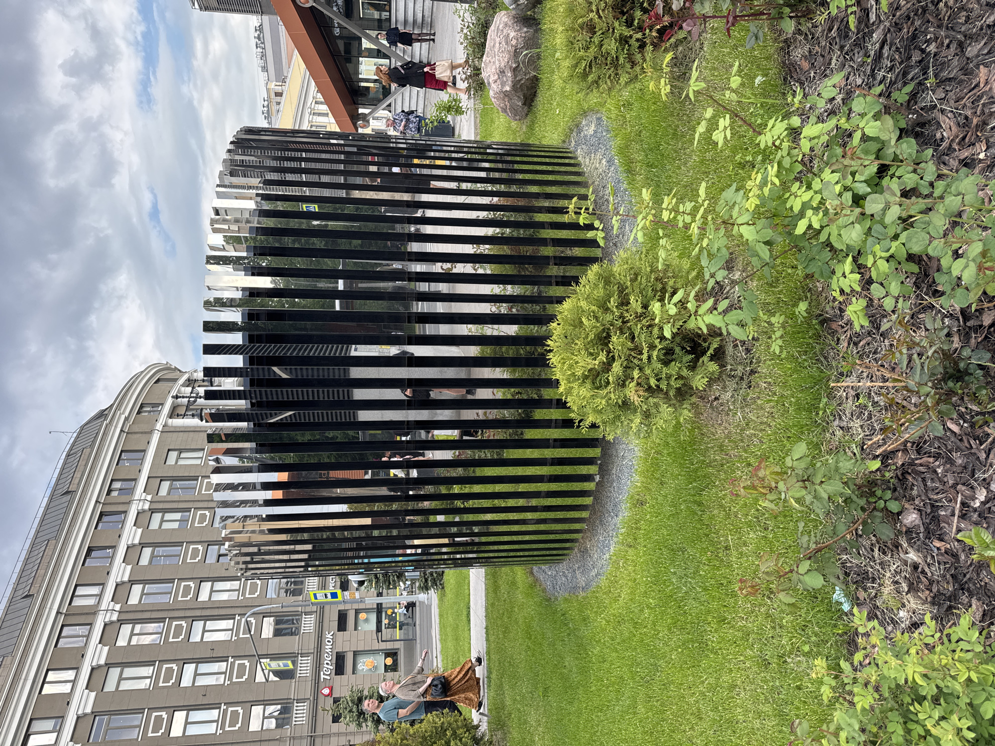 This image captures a vibrant urban scene in Saint Petersburg, Russia, featuring modern architecture, public green spaces, and pedestrians under a partly cloudy sky. The foreground is dominated by a large, curved, reflective art installation or screen composed of vertical black slats. This structure acts as a series of mirrors, distorting and reflecting the surrounding environment, including nearby buildings and the sky.
To the right of the reflective structure is a well-maintained green lawn interspersed with various shrubs and bushes, some with darker foliage and others with lighter green leaves. A large, grey boulder sits among the greenery. Parts of the lawn are bordered by areas covered in wood mulch.
In the mid-ground, a man with light hair wearing a light blue shirt is seen walking or standing, gesturing slightly with his right hand. Beside him, a woman with light hair, dressed in a brown patterned skirt and dark top, carries a small black handbag. Further back, near the entrance of a building, other people are visible, including a woman in a dark dress and white top, and another person in a dark coat and red skirt.
Behind the green space and reflective structure, a multi-story building with a distinctive curved facade and rectangular windows dominates the left side of the frame. This building exhibits classical architectural elements like cornices and pilasters. On its ground floor, a sign with the word "Теремок" (Teremok), a well-known Russian restaurant chain, is clearly visible, confirming the Saint Petersburg context. Another building with a brown-orange overhang is partially visible through the reflections in the background.
The scene takes place during the daytime, with soft, diffused light suggesting an overcast or partly cloudy sky. A pedestrian crosswalk sign can be seen reflected or directly through the slats of the art installation. The overall impression is one of a contemporary city blending natural elements with modern and historic architecture.
