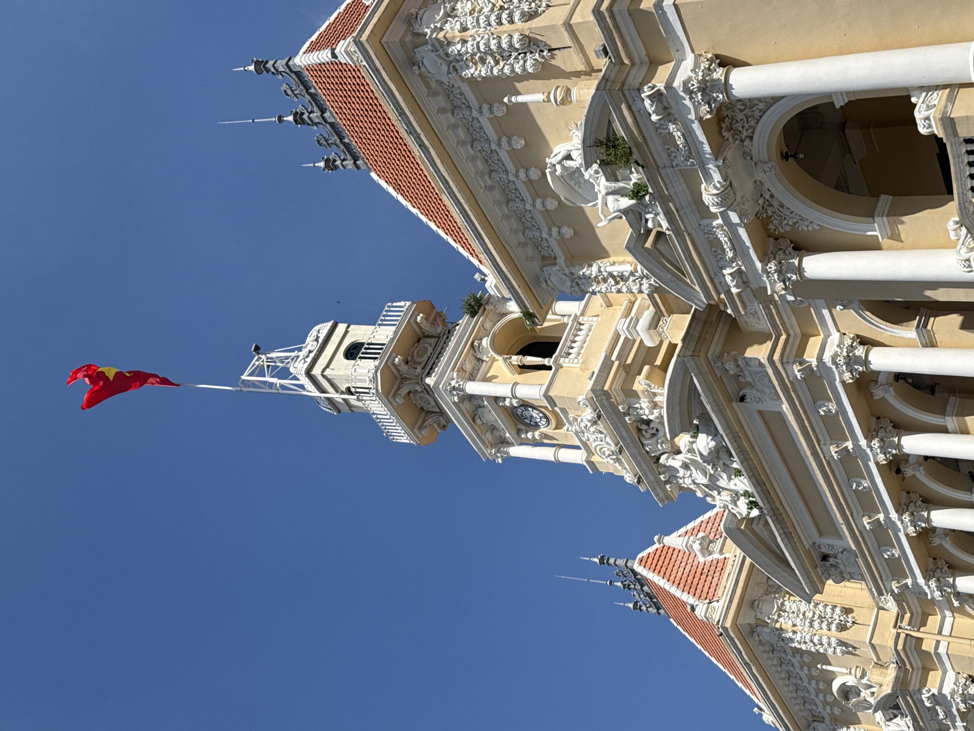 The image shows the facade of the Ho Chi Minh City Hall, located in District 1 of Ho Chi Minh City, Vietnam. The building is a French colonial-era structure with ornate architectural details, including sculptures, columns, and a red-tiled roof. A Vietnamese flag flies from a flagpole on top of the central clock tower. The sky is clear and blue.