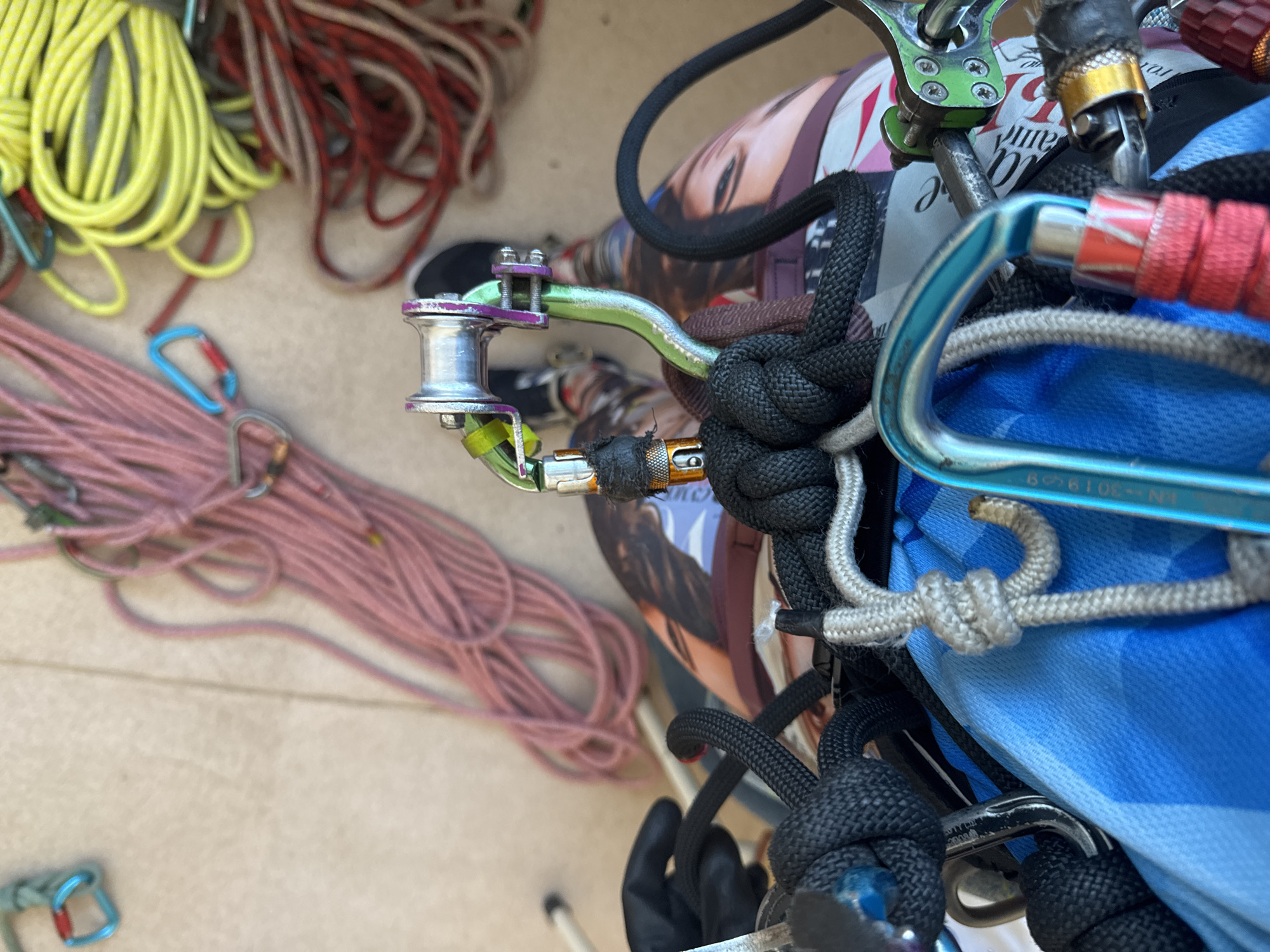 The image depicts a collection of climbing gear, including ropes of various colors (yellow, red, pink) and numerous carabiners, some attached to a complex arrangement of knotted ropes. A pulley system with a silver wheel and lime green arm is also visible. The background is a mix of indistinct objects and surfaces, including what appears to be a magazine with a woman's face on the cover. The location (Usolye, Russia) is not discernible from the visual information provided.