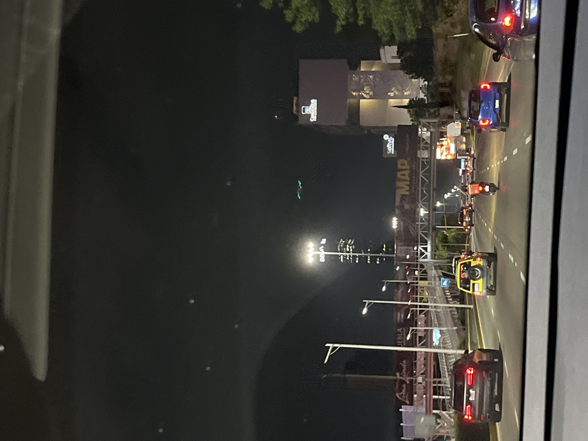The image displays a nighttime urban or highway scene, viewed from inside a vehicle looking out onto a multi-lane road. The perspective is slightly elevated, and a portion of the vehicle's interior frame is visible on the far right.
Numerous cars and at least two motorcycles are on the road, with their headlights and taillights illuminating the scene. Vehicles include a blue sedan, a dark-colored SUV in the foreground, and a distinctive yellow off-road SUV (resembling a Jeep) with a rear-mounted spare tire. Streetlights brightly illuminate the road and surrounding area.
Along the roadside, there are various buildings and large sign structures. A prominent billboard-like gantry displays the text "MAP" in large white letters, with "PUEBLA" visible underneath. Another illuminated sign further down reads "Softys". In the background, additional buildings are lit up, including one featuring the "Cinemex" logo and another with what appears to be the "BBVA" logo. Some green foliage from trees can be seen above the buildings. The sky is dark, clearly indicating the time of day is night.
The activity depicted is active traffic on a major road or highway. Small white dots scattered across the dark sky areas may be reflections or lens flares. The scene is located in San Bernardino Tlaxcalancingo, Mexico.