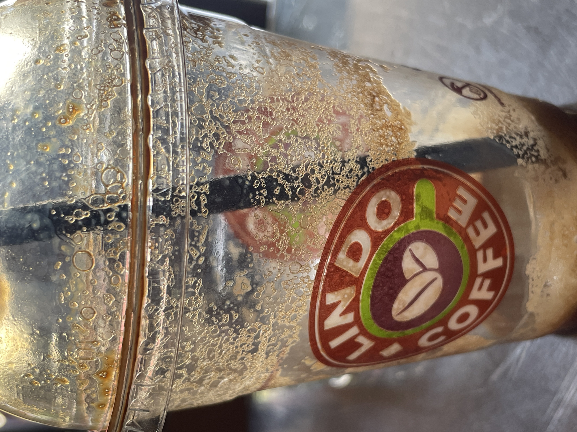 A close-up, slightly out-of-focus shot shows the interior of a used, clear plastic coffee cup with a domed lid. The inside of the cup and lid are coated with remnants of a brown beverage and small bubbles. A black straw is visible, extending through the lid. A circular logo with the text "LINDO COFFEE" and an image of coffee beans is prominently displayed on the side of the cup. The lighting creates reflections and highlights on the plastic. The background is blurred and appears to be a dark, possibly metallic surface. The scene is indoors.