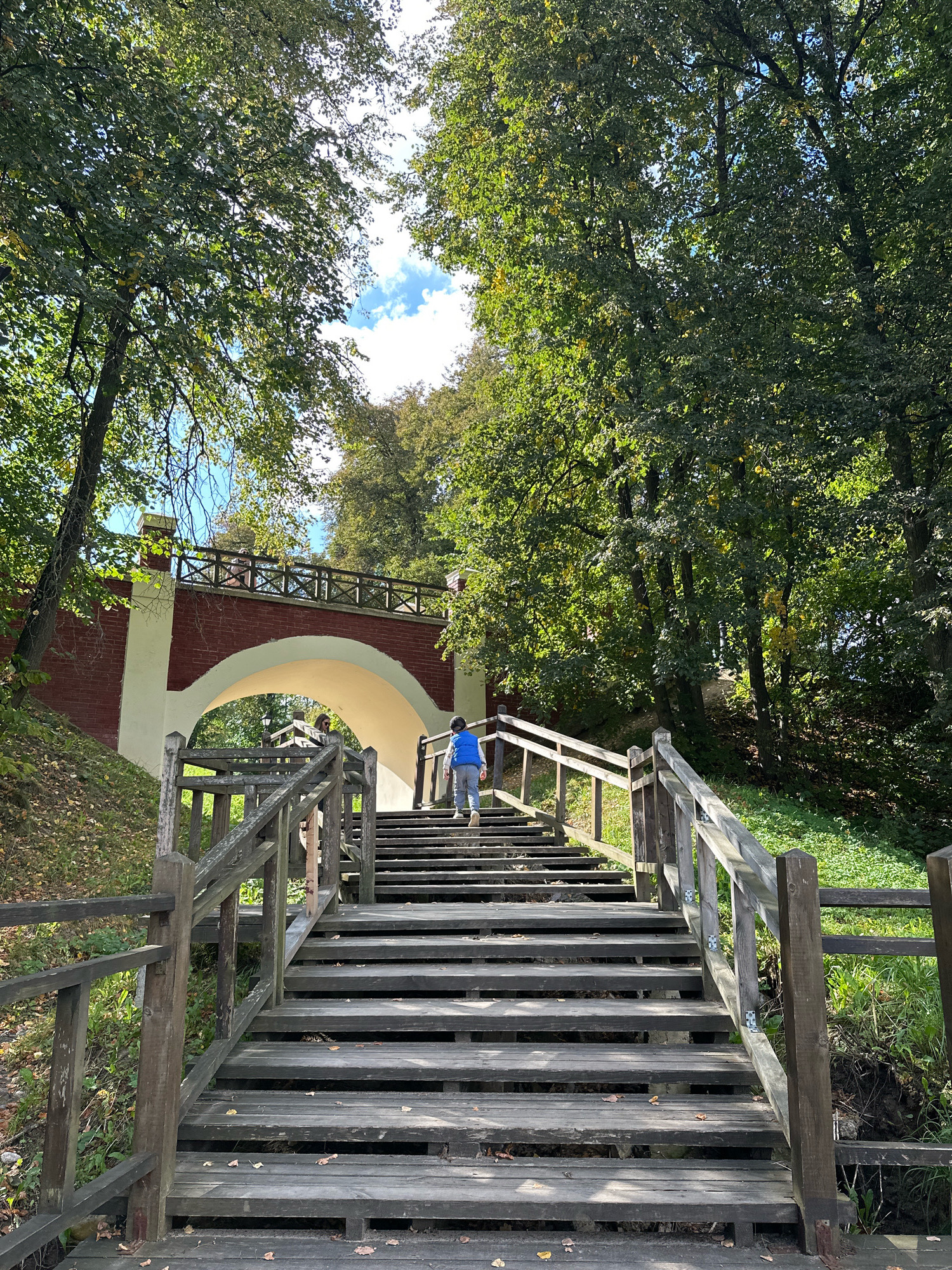 The image depicts a wooden staircase ascending towards a brick bridge with an arched opening. The staircase is made of weathered wood planks and features a wooden railing. A young person wearing a blue vest and grey pants is walking up the stairs. The bridge, constructed from red bricks and white stucco, spans over a path or natural terrain. The surroundings are lush with green trees and foliage, suggesting an outdoor, possibly park-like setting. The sky is visible through the leaves, with some clouds present. The scene appears to be filmed from a low angle, looking up the stairs and towards the bridge. The environment suggests a sunny day. The specific location in Minsk, Belarus is not identifiable from the visual information provided.