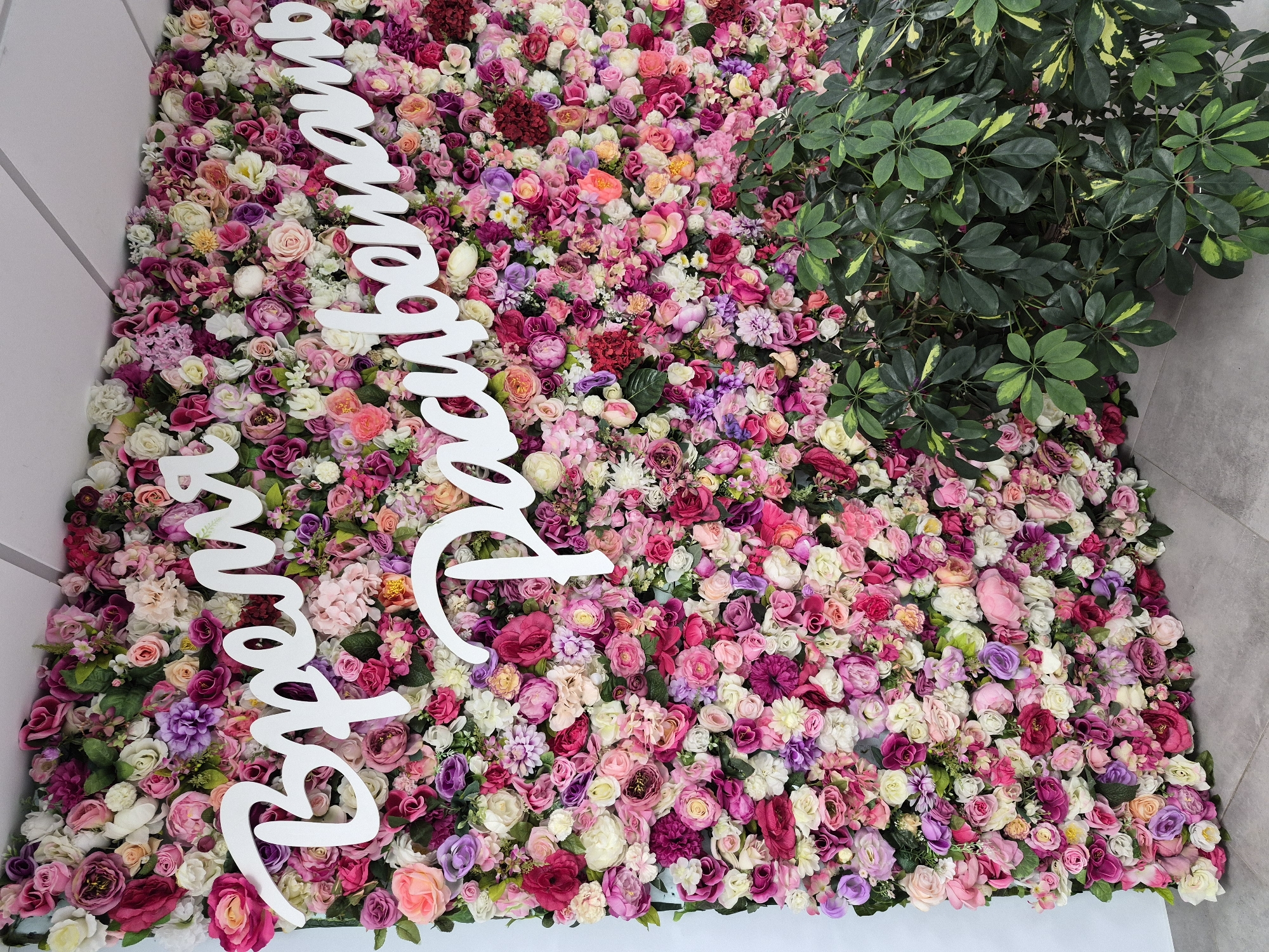 A vibrant floral wall serves as a backdrop for white, cursive lettering. The wall is densely packed with artificial flowers in various shades of pink, purple, and white, creating a rich tapestry of color and texture. The white letters, which spell out "Время расцветать" (Vremya rastsvetat' - "Time to bloom"), are prominently displayed across the floral arrangement. In the foreground, a lush green plant with broad leaves partially obscures the lower section of the floral wall. The floor appears to be made of light grey tiles. The scene is indoors, with a white ceiling visible at the top. This setup is likely a decorative element in a public space, such as a shopping mall, event venue, or a beauty salon in Yekaterinburg, Russia, designed to evoke a sense of beauty and positivity.