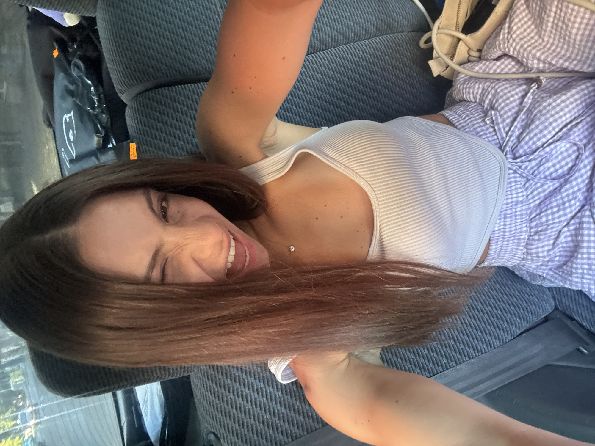 A young woman is photographed from the chest up, winking and smiling widely. She has long brown hair, a white ribbed tank top, and light purple checkered pants. She is sitting in the back seat of a car, with car seats visible behind her. Her right arm is extended, likely holding the camera for the selfie. A light-colored handbag rests beside her on the seat. The scene is an interior of a vehicle, with hints of the outside through the window suggesting it is daytime.