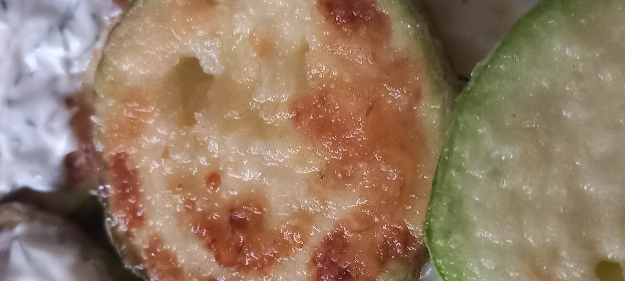 A macro photograph depicts two slices of cooked produce. The dominant upper slice is circular, presenting a glossy, golden-brown, and irregularly textured surface with localized darker reddish-brown charring or caramelization. A small, central void is visible within this slice. Positioned below and partially obscured is the curved perimeter of a second, lighter green slice with a distinct outer green rind and a paler interior. The background is highly blurred, presenting a light-colored, indeterminate surface. The depicted scene is located in Kryvyi Rih, Ukraine.