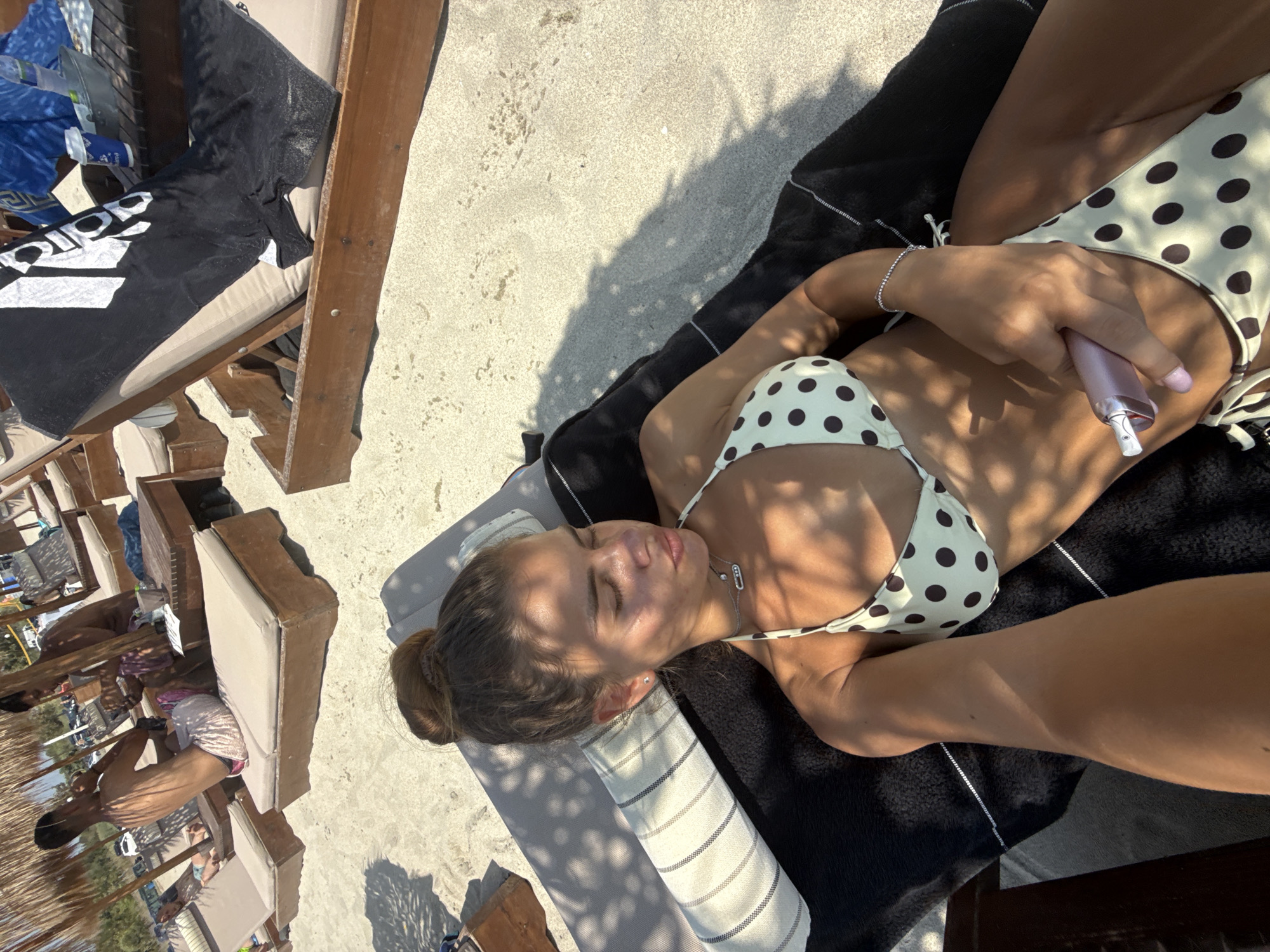 In Katerini Municipality, Greece, a young woman with her hair in a bun is sunbathing on a beach lounger. She is wearing a polka dot bikini and has her eyes closed, seemingly enjoying the sun. Her right hand is resting on her lower abdomen, holding a pink cylindrical object. The lounger is covered with a black towel and a striped pillow. The scene is set on a sandy beach, with other loungers and umbrellas visible in the background, suggesting a popular tourist spot. Sunlight filters through overhead foliage, casting dappled shadows on the sand and the woman. Other individuals can be seen in the distance, further populating the beach.