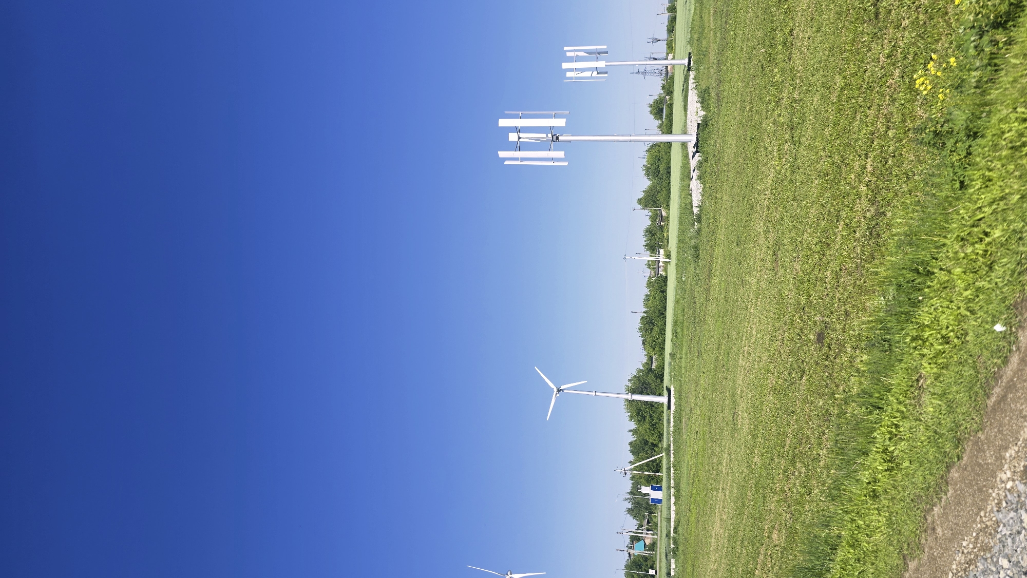 The image depicts a clear, bright day with a vast expanse of deep blue sky. In the foreground, a green, grassy field stretches across the frame, with a gravelly path visible on the lower left. In the mid-ground, several wind turbines are positioned within the field. To the left, a traditional three-bladed horizontal axis wind turbine stands tall. Further to the right, two smaller vertical axis wind turbines with multiple blades are prominent. A line of green trees forms a backdrop across the mid-ground, behind which a few buildings and power lines are discernible, suggesting a rural or semi-rural setting on the outskirts of a settlement. The overall scene conveys an impression of open space and renewable energy infrastructure. The location is confirmed to be the city of Актюбинский, Russia.