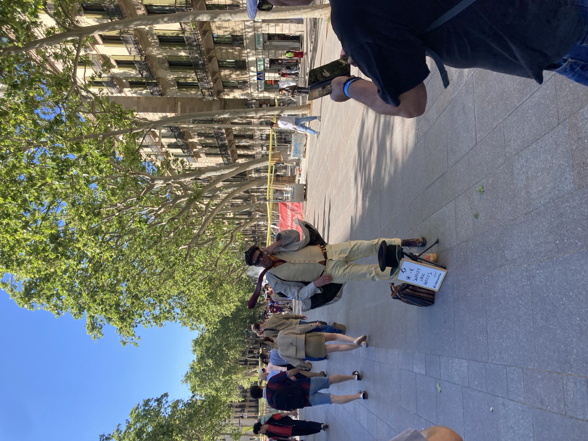 A street scene in Unknown, Unknown features a sidewalk with pedestrians walking in both directions. On the right side of the image, a man in a black shirt and blue jeans is holding a phone and appears to be taking a picture of a street performer. The performer is wearing a beige suit with a black cape and hat and stands in the center of the sidewalk with a sign that reads "Support Local Artists" next to a hat and bag. A row of tall buildings and trees lines the left side of the image. The weather is sunny.