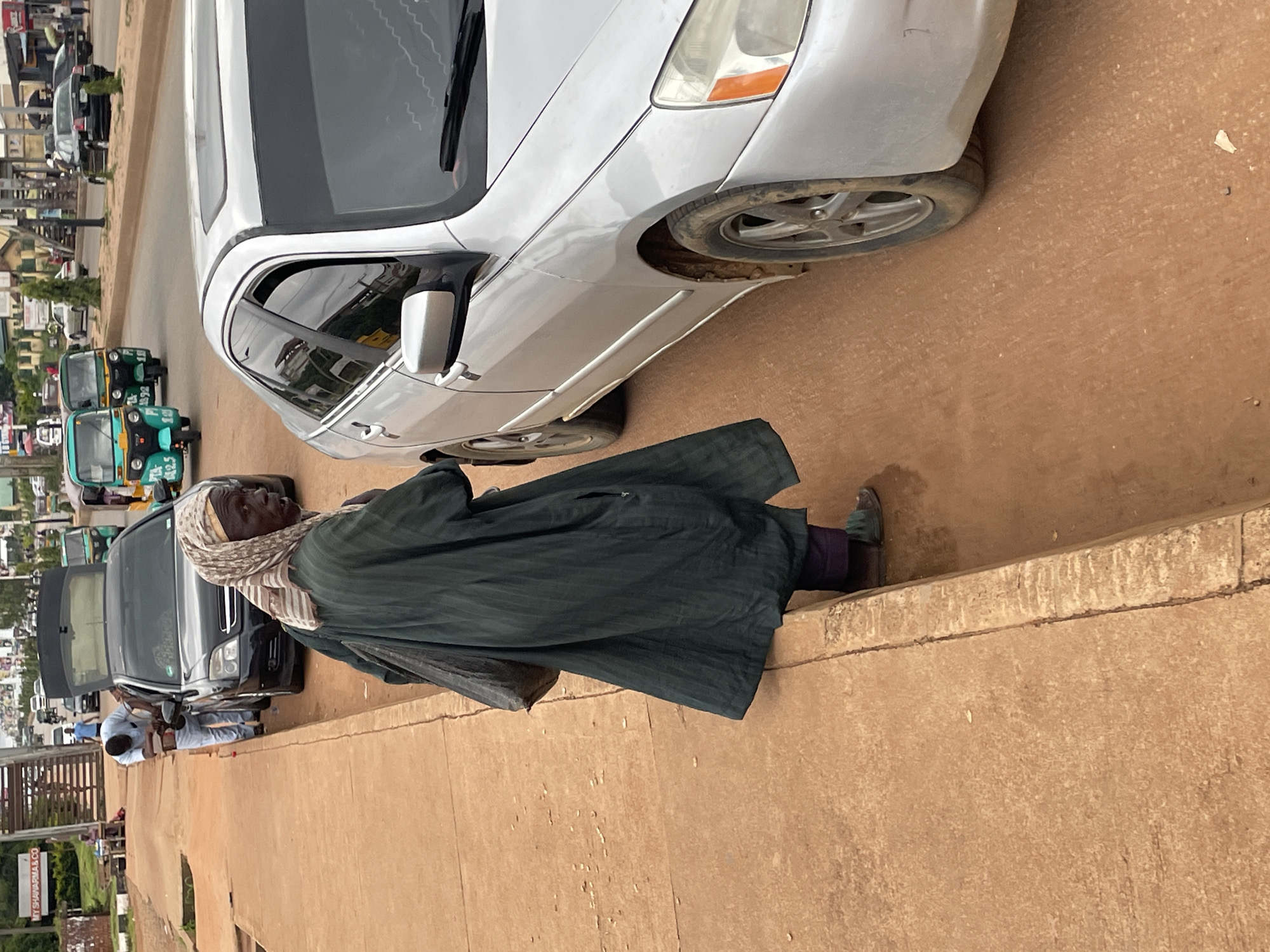 The image depicts a street scene in Jos, Nigeria, with a focus on a woman standing on a sidewalk next to a silver sedan. The woman, who appears elderly, is wearing a dark green robe and a patterned headscarf. She is carrying a dark bag over her shoulder. To her left, a black car with its trunk open is parked, and a person in a light blue shirt and light-colored trousers is attending to it. Behind the silver sedan, a teal and yellow motorized rickshaw is visible in the street. The sidewalk is made of concrete, and the street is paved with asphalt and appears somewhat dirty. In the background, there are buildings, other vehicles, and pedestrians, suggesting a moderately busy urban environment. The overall lighting indicates it is daytime.