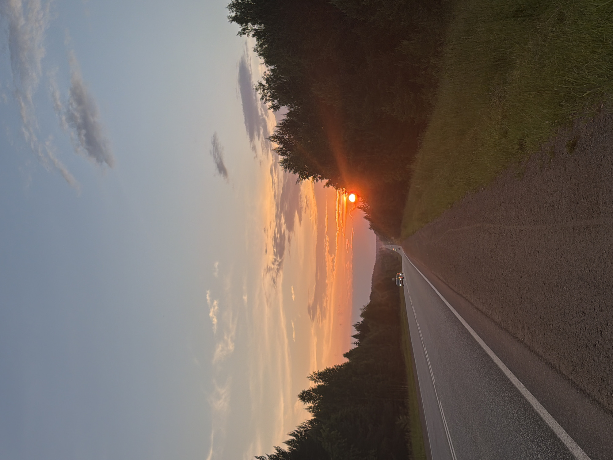 The image depicts a sunset scene on a straight, asphalt road, likely in Chaikovsky, Russia. The road runs through a dense forest on both sides, with the setting sun casting a warm orange and yellow glow across the sky. The sun is visible on the right side of the image, partially obscured by trees, creating lens flare. A few cars are visible on the road, heading away from the viewer. The sky is a gradient of pale blue to orange, with scattered clouds. The overall impression is one of a serene, rural landscape at dusk.