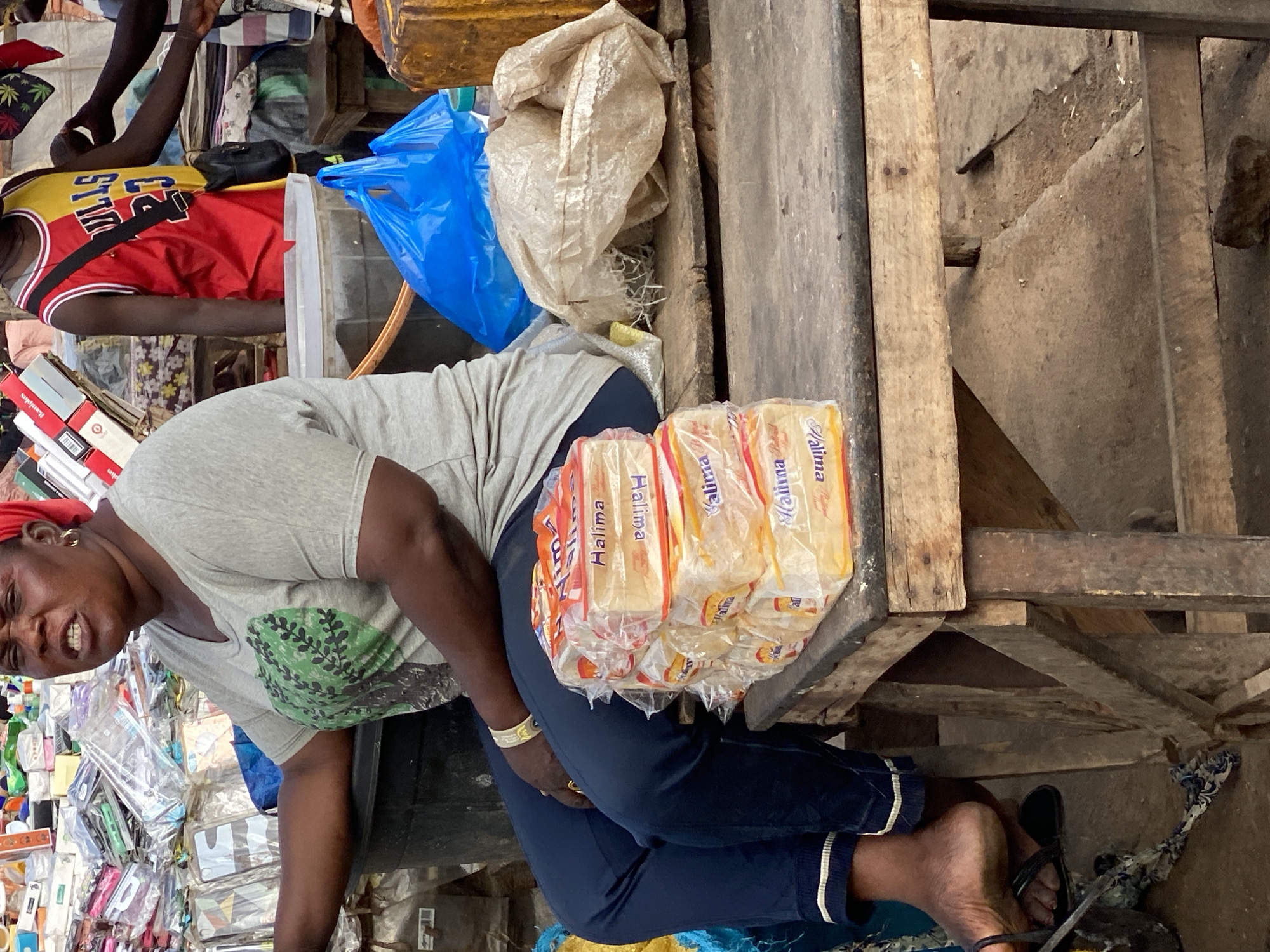 The image depicts a street scene in Sabon Tasha, Nigeria, likely in a market setting. The primary subject is a woman, seen from the chest down, sitting on the ground. She is wearing a grey t-shirt with a green tree graphic, dark blue pants with white stripes on the cuffs, and a red headscarf. She has a gold bracelet on her left wrist and is wearing a gold ring on her right hand. Her facial expression suggests strain or exertion, with her mouth open and teeth bared.
Beside her are several loaves of bread, packaged in clear plastic with orange and yellow labels that read "Halima." These appear to be the main items for sale. In the background, there are various other objects and people, though they are less distinct. To the left, a person wearing a red and yellow basketball jersey can be seen. Various goods, including plastic bags and boxes, are piled around the scene, indicating a busy marketplace. The overall impression is one of a vendor at work in a crowded, somewhat disorganized environment. The lighting suggests it is daytime, and there are no clear indicators of the weather. The presence of diverse goods and the general atmosphere point to a typical Nigerian market.