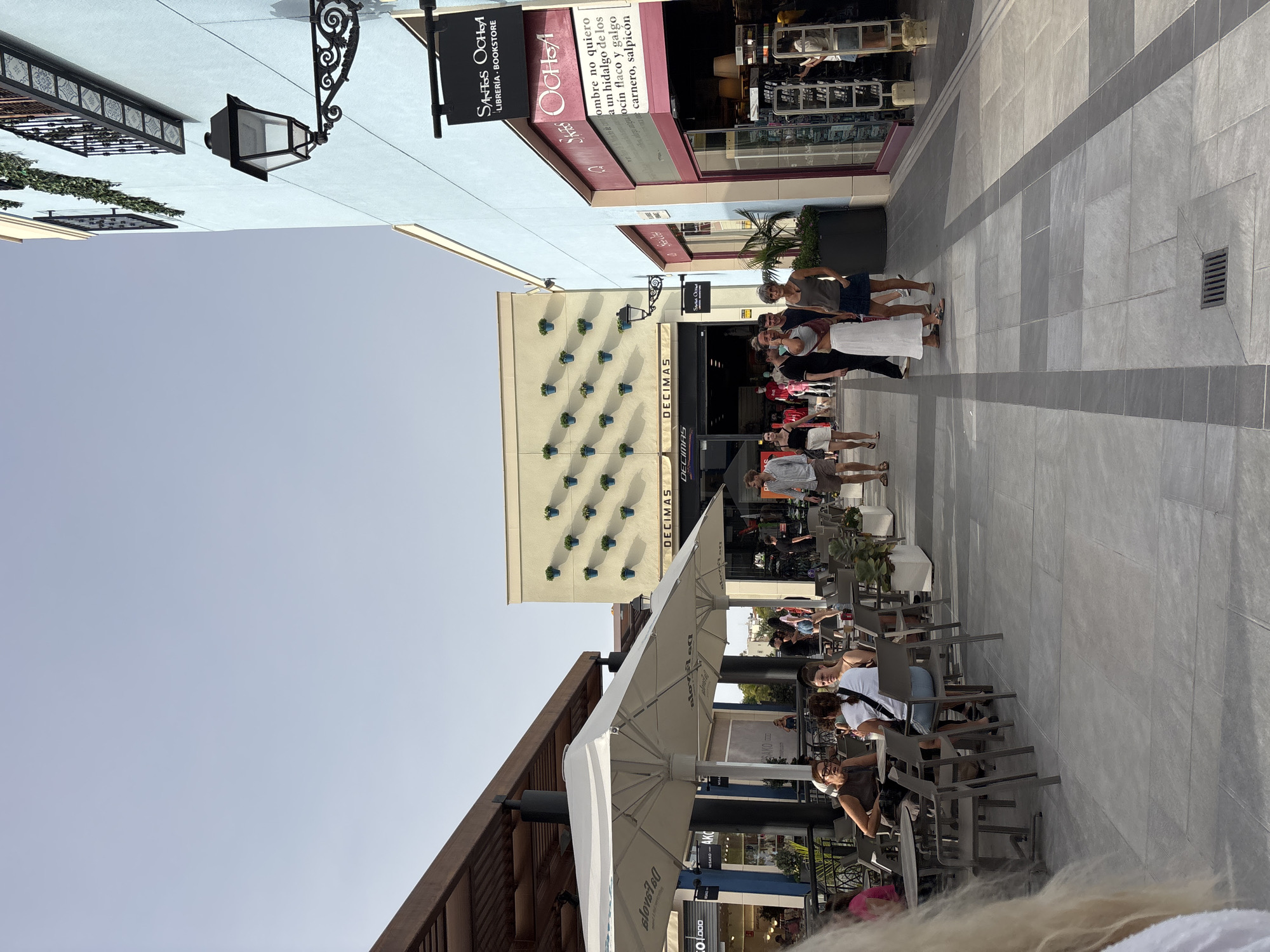 The image shows an outdoor pedestrian walkway in Orihuela, Spain, under a clear sky. On the left, a covered seating area with tables and umbrellas is occupied by several people, some seated and others standing. The architecture features light-colored walls and wooden overhangs. To the right, a pale blue building with a black streetlamp and a sign reading "SANTOS OCHOA LIBRERÍA • BOOKSTORE" is prominent. Further down the walkway, a cream-colored building is decorated with rows of blue potted plants on its facade. Other people are walking along the walkway, and shop fronts are visible. The ground is paved with large, light-colored tiles arranged in a grid pattern with darker borders.