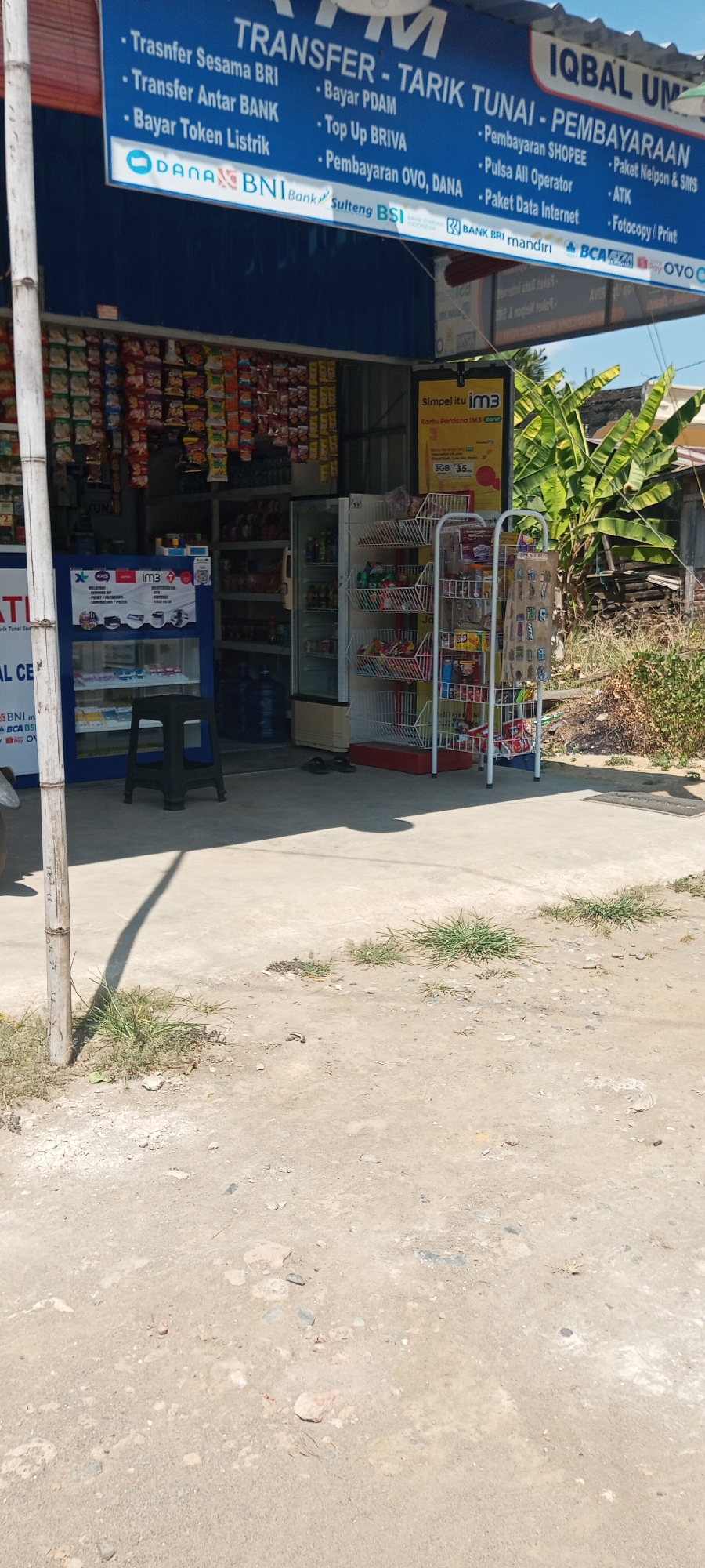 The image depicts the exterior of a small convenience store, likely in Lempa, Indonesia, identified by a sign that reads "IQBAL UM". The store displays a blue awning with text listing various financial services such as "TRANSFER SESAMA BRI", "Bayar PDAM", "Top Up BRIVA", and "Pembayaran OVO, DANA". Below this, logos for DANA, BNI Bank, and Sulteng BSI are visible, indicating financial transaction capabilities.
The storefront features shelves stocked with numerous packaged goods, including snacks and drinks. To the right, a white metal rack holds more products. A refrigerator unit is also present within the store. Outside the store, a black plastic stool is placed on the concrete ground. A sign for "im3" (a mobile carrier) is prominently displayed. The ground in front of the store is unpaved, with patches of dry grass and scattered rocks. The background reveals a vegetated area with banana trees and some discarded wooden items. The overall scene suggests a busy local retail establishment offering a range of goods and services.