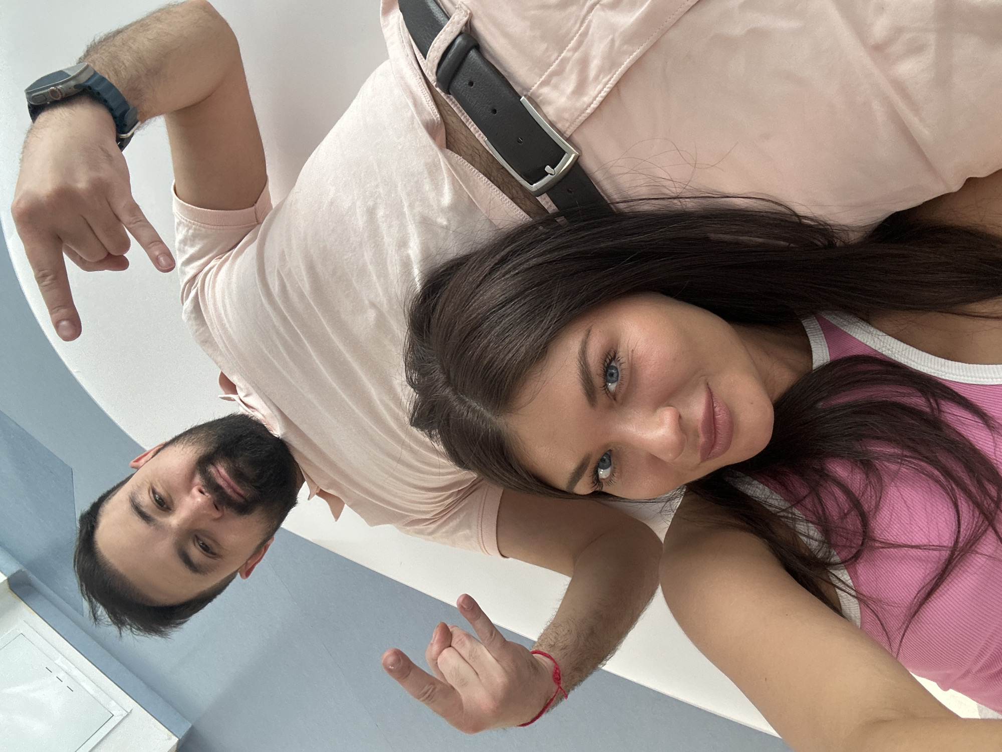 A selfie captures a man and a woman in Khimki, Russia, from a rotated perspective. The man on the left has a beard and wears a light pink shirt and a smartwatch on his left wrist. He is gesturing downwards with his right hand. A black leather belt with a silver buckle is visible on his shirt. The woman on the right has long, dark hair and wears a pink tank top with white trim. She is gesturing with her left hand, displaying the "rock on" sign. Her wrist is adorned with a red bracelet. The background appears to be a white wall with a light blue accent.