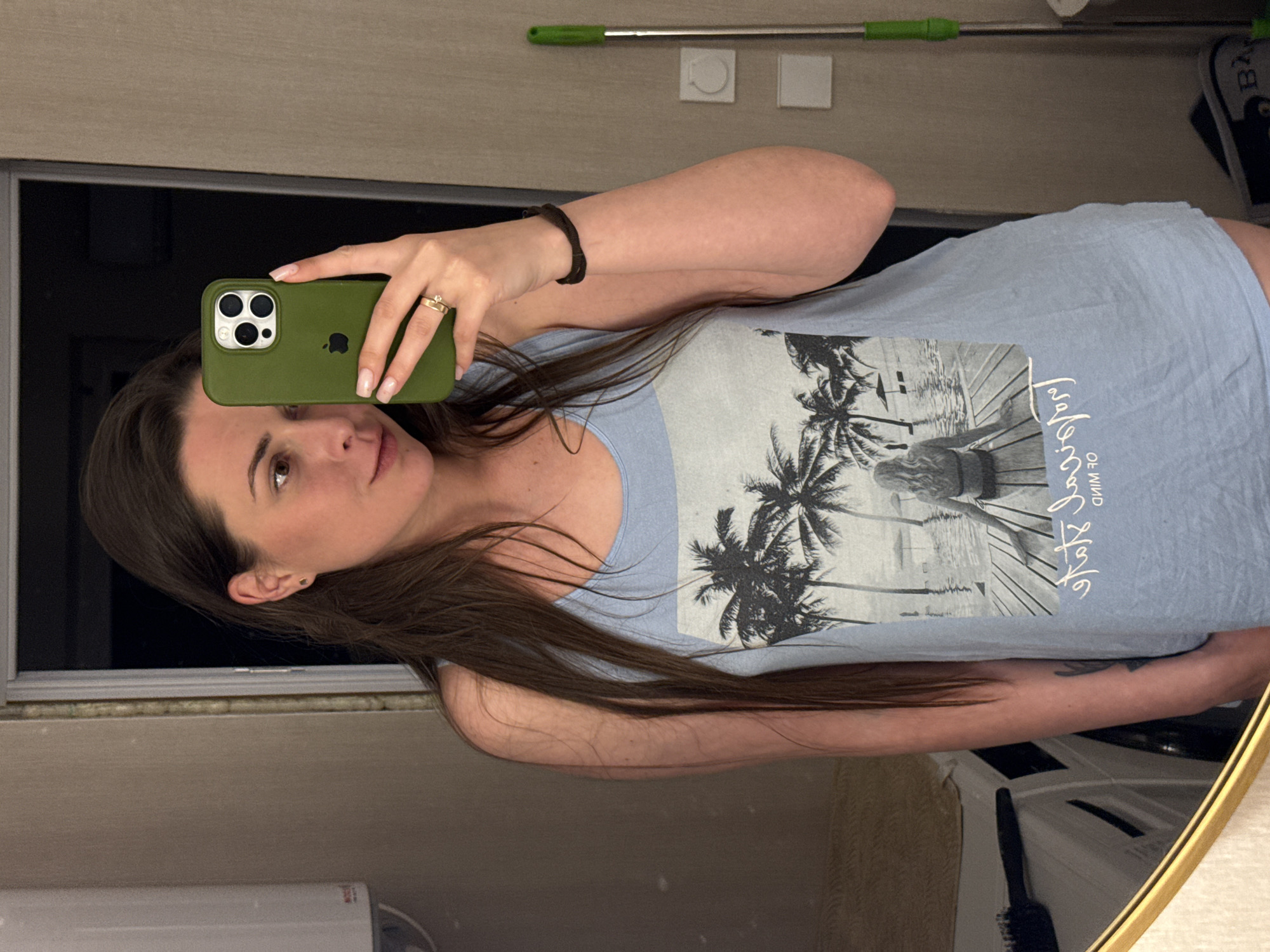 A young woman with long brown hair tied back is taking a selfie in a mirror. She is wearing a light blue tank top with a graphic print of palm trees and a woman sitting on a wooden deck. She is holding a green iPhone in her right hand. She has a ring on her left ring finger and a dark bracelet on her left wrist. The background appears to be a room with a door, a mirror, and a floor mop. The woman is looking at the camera and smiling slightly. There is a water heater on the left side of the image and a toothbrush on a counter reflected in the mirror on the lower left.