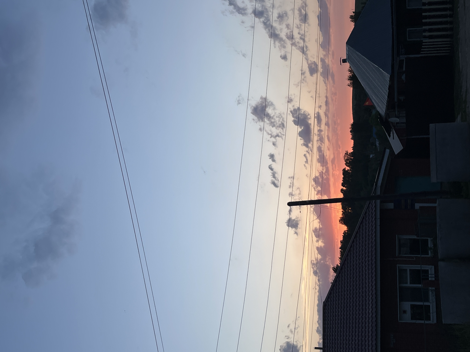 The image depicts a sky with a gradient of colors transitioning from blue on the left to orange and pink near the horizon on the right. Scattered clouds are present throughout the sky. Multiple power lines run horizontally across the image. On the right side, there are houses with tiled roofs and dark silhouettes against the vibrant sky. The location is Фоки, Russia.