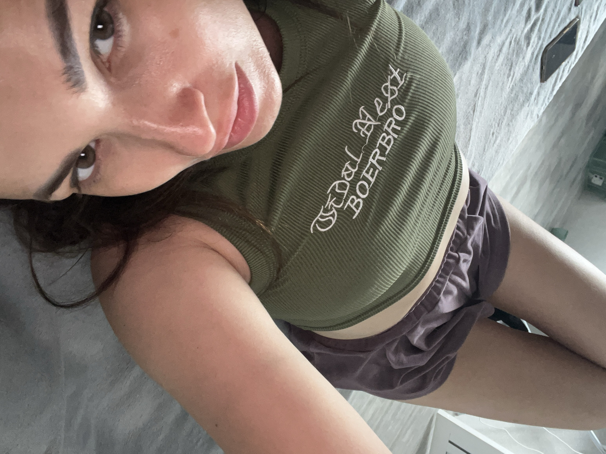 A woman is lying on her back on a bed, looking up at the camera. She is wearing a green ribbed tank top with white text that reads "Dal Boerbro". She is also wearing a pair of purple shorts. Her legs are crossed. A smartphone is visible on the bed to her left. The room is sparsely furnished and appears to be a bedroom. The lighting is natural, coming from a window or overhead light.