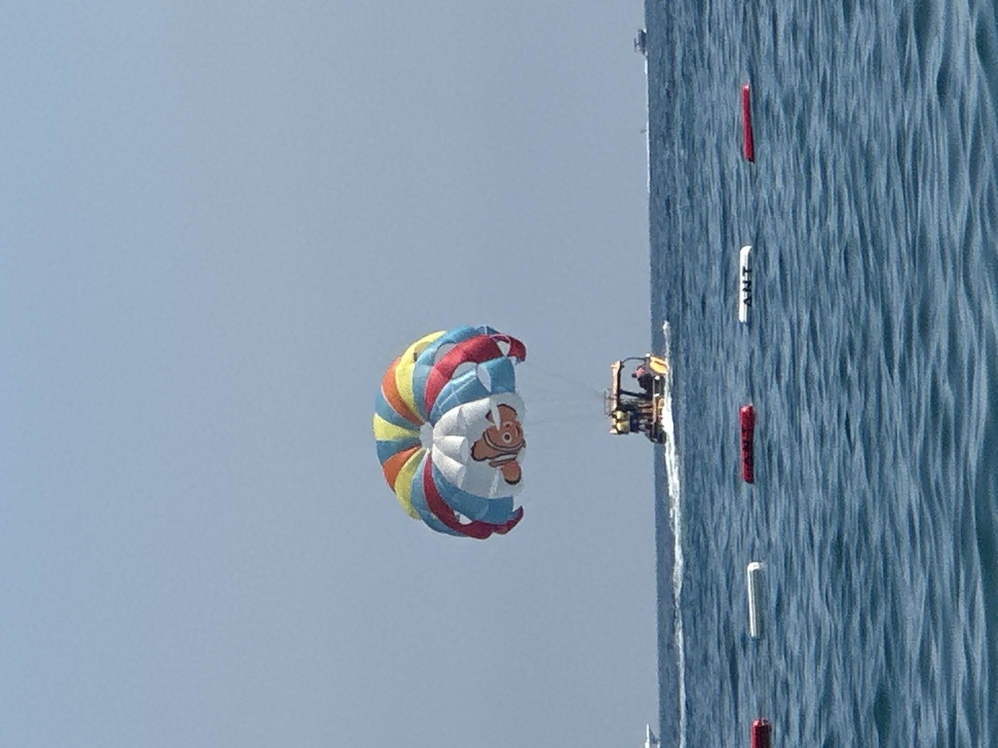 In the city of Boreti, Montenegro, on a clear, bright day, a parasail floats in the sky, tethered to a boat moving through the calm, blue sea. The parasail is brightly colored in red, yellow, and blue segments and features a cartoon image of a fish, resembling Nemo from the animated film "Finding Nemo." A small boat is attached to the parasail, carrying two individuals who appear to be operating the vessel. Several buoys are visible floating on the water's surface, some red and one white with black lettering that reads "ANT". In the distance, other boats are visible on the horizon. The water is slightly rippled, indicating a gentle breeze.