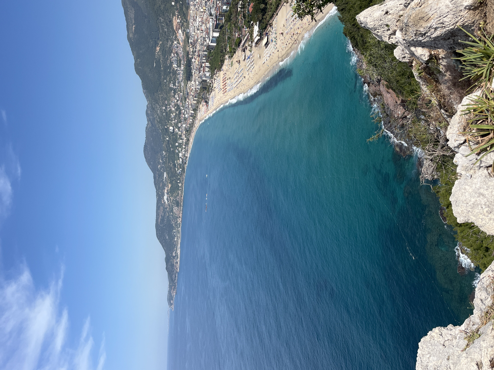 The image depicts a high-angle view of a coastal scene in Alanya, Turkey. A long sandy beach is lined with numerous closely spaced sun umbrellas, suggesting a popular tourist destination. Behind the beach, a densely populated town extends up the slopes of a mountain. The sea transitions from a turquoise color near the shore to a deeper blue further out. The foreground features a rocky cliff edge with sparse vegetation. The sky is clear blue with only a few thin clouds.