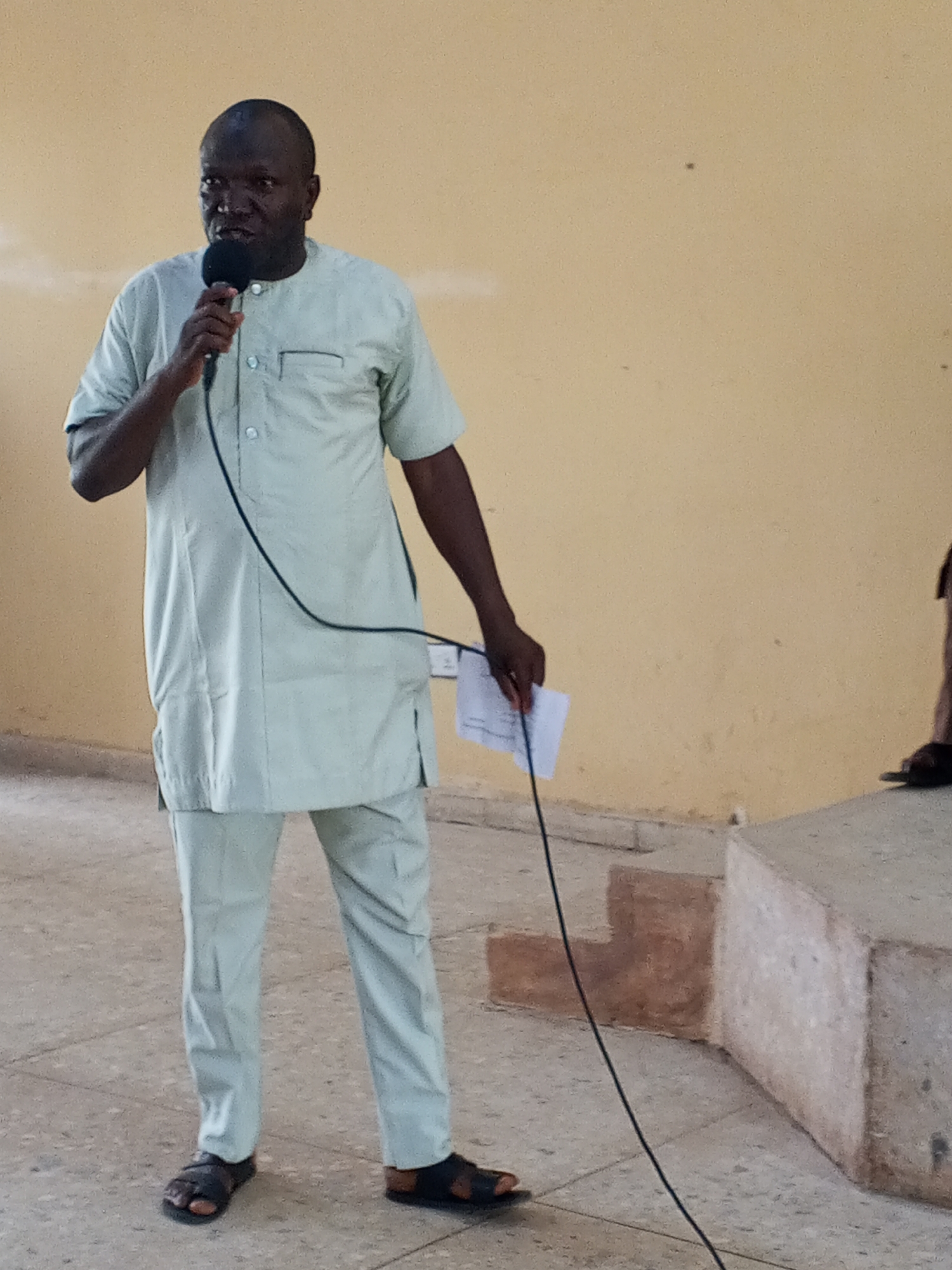 A man stands on a concrete floor in front of a pale yellow wall, holding a microphone in his left hand and a piece of paper in his right hand. He is wearing a light green traditional outfit and sandals. To his right is a concrete structure that is slightly elevated. The location is Unknown, Unknown.