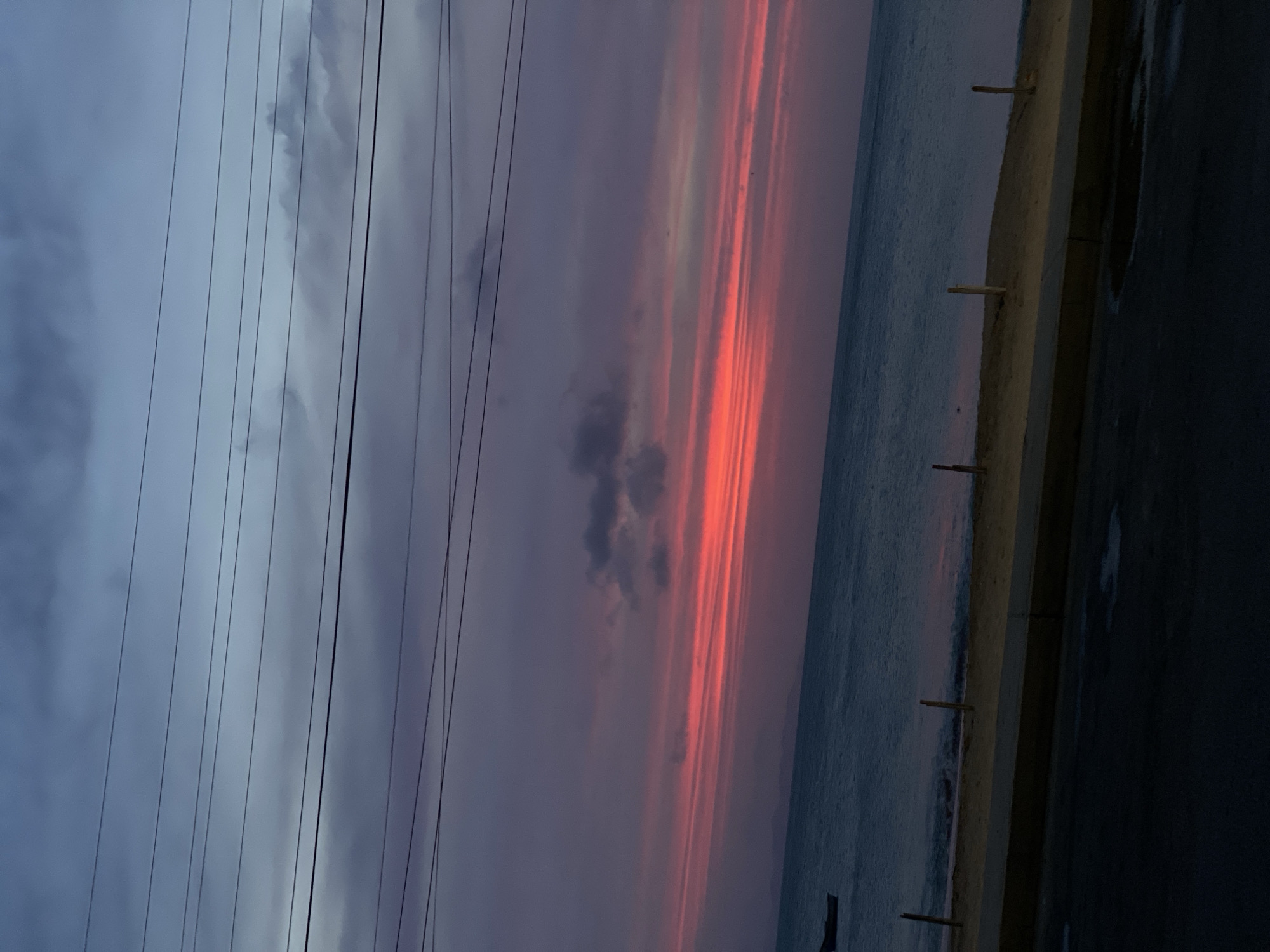 Here is a forensic description of the image:
The image depicts a coastal scene, possibly at sunset, in Juan Griego, Venezuela. A beach is visible alongside the ocean. The sky shows a dramatic color gradient, shifting from greyish-blue with visible power lines in the left portion of the image, to shades of pink and orange near the center and right. The sun, although not visible directly, casts an orange/red glow through the clouds. The presence of dark clouds in the center of the image contributes to the atmospheric contrast. On the right, there's a paved area, likely a road or walkway, adjacent to the beach. Wooden pilings are planted at regular intervals along the edge of the shore. The perspective of the image is slightly tilted. No people are readily visible in the scene.