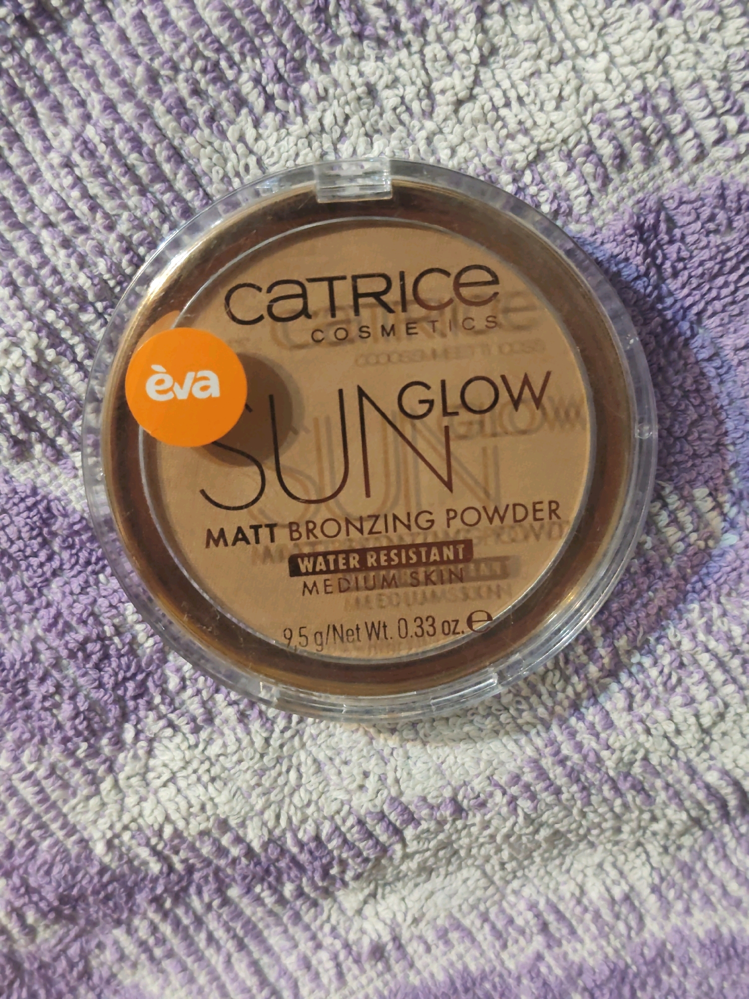 A close-up overhead shot shows a round, clear plastic compact of Catrice Sun Glow Matt Bronzing Powder with a bronze-colored powder inside. The compact is placed on a textured purple and white towel. The brand name "CATRICE" is printed in black on the top half of the powder, followed by "COSMETICS" in smaller letters. The words "SUN GLOW" are written largely across the center of the powder in a dark brown font. Below this, the text "MATT BRONZING POWDER" is visible, with "WATER RESISTANT MEDIUM SKIN" and "9.5 g/Net Wt. 0.33 oz. e" printed below in smaller fonts. An orange circular sticker with the word "eva" in white lettering is affixed to the left side of the compact. The background is a soft focus of the purple and white striped towel.