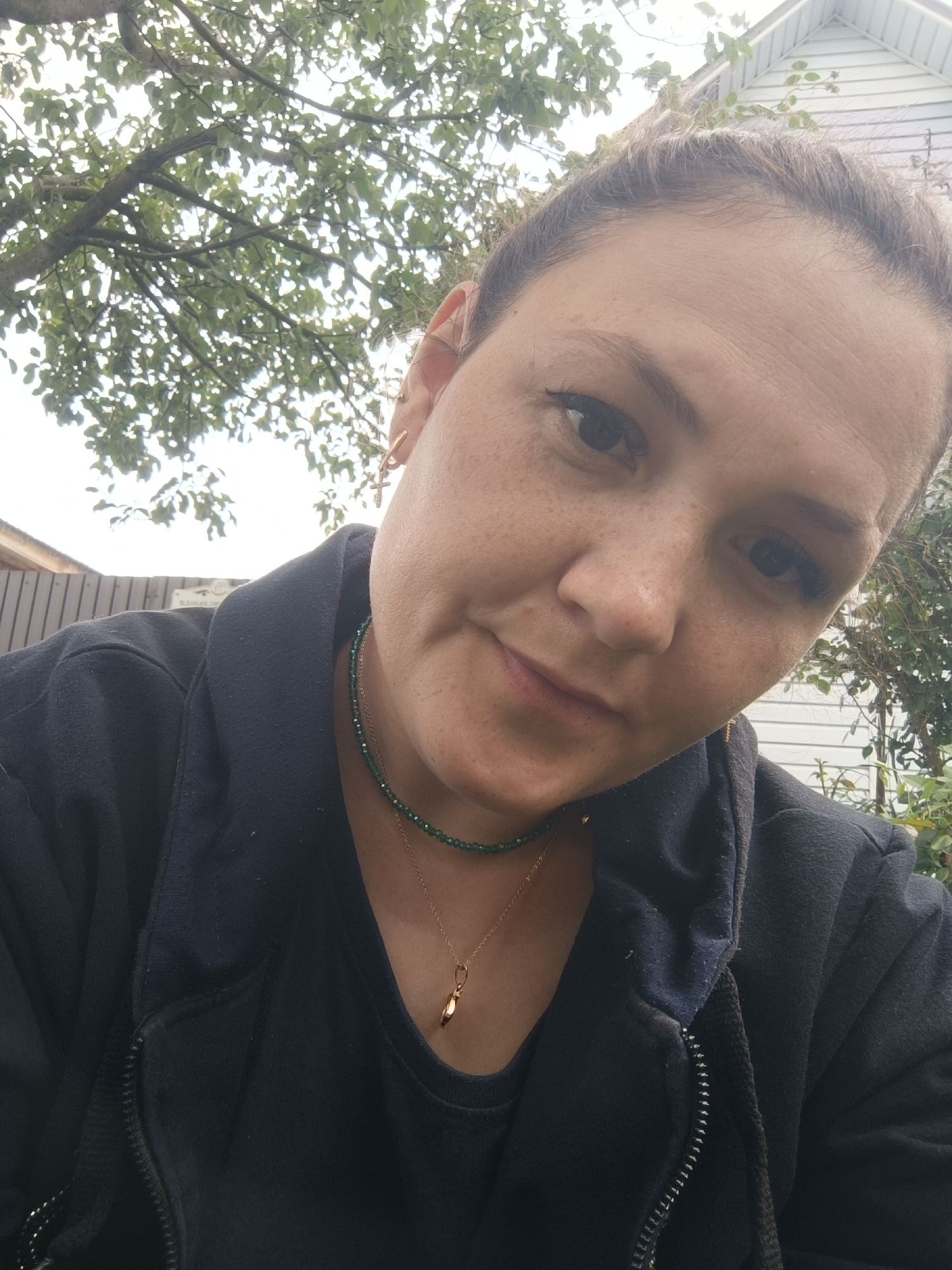 A medium shot photograph shows a woman with light brown hair pulled back, wearing a dark blue hooded jacket over a black shirt. She has freckles on her face and is wearing a thin green necklace with a gold pendant. She also has a gold earring in her visible ear. The background consists of foliage from a tree, a wooden fence, and a white building. The woman is looking towards the camera with a slight smile. The scene appears to be outdoors, possibly in a residential area. There is no information in the image to indicate the location is the city of Karsha, Russia.