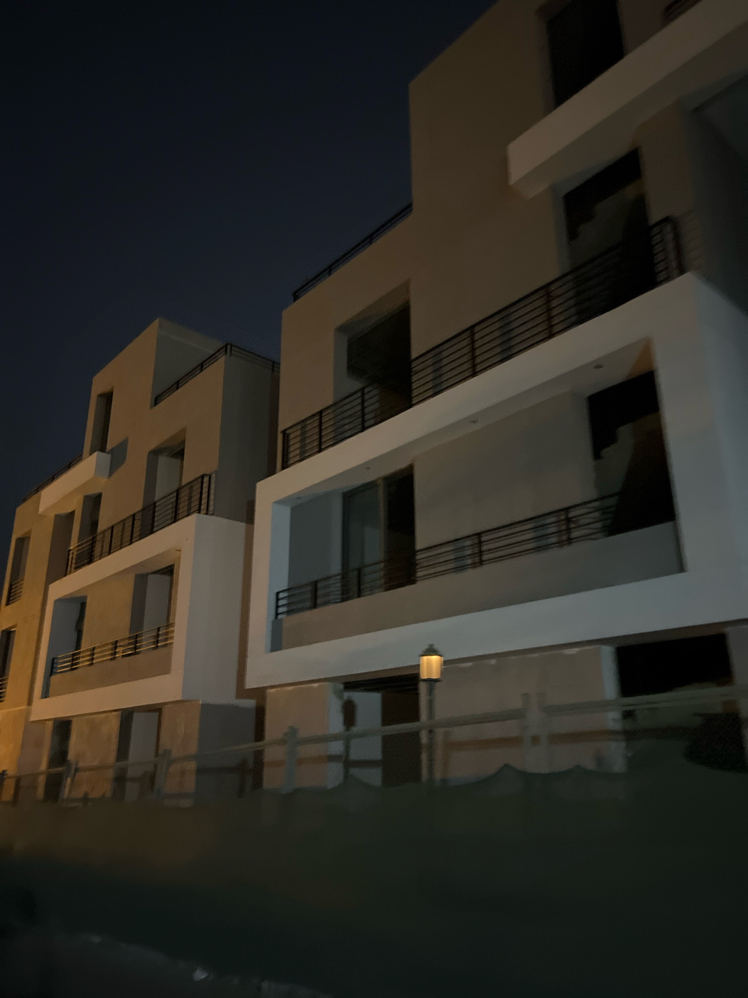 The image depicts the exterior of a modern, multi-story building complex at night. The architecture features clean lines, concrete facades in beige and white, and dark metal railings on balconies. The building is partially illuminated by artificial lights, casting shadows and highlighting its geometric forms. The sky is a deep, dark blue, indicating nighttime. A street lamp with a warm, yellow glow is visible in the foreground, along with a wooden fence. The overall impression is one of contemporary construction in an urban or suburban setting. The context provided places this scene in Sharm El Sheikh, Egypt, suggesting it could be a residential or commercial development in that city. There are no people visible in the image.