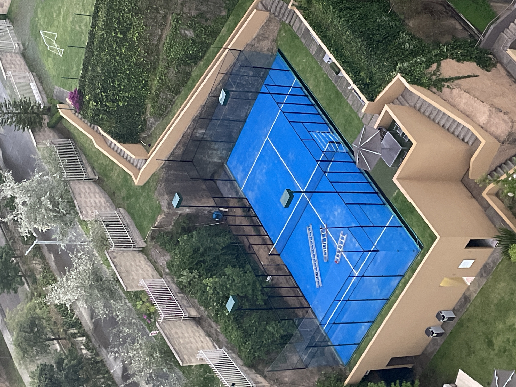 The image presents an aerial view of a paddle tennis court, located in Huixquilucan de Degollado, Mexico. The court is painted blue with white lines marking the boundaries. It is enclosed by glass walls and black metal fencing. There are multiple ladders on the court. The court is surrounded by greenery, including trees, bushes, and grass. To the left of the court are stairs with white metal railings, and to the right is a multi-story building with an outdoor seating area and staircase. The buildings appear to be residential. There is a green lawn with a soccer goal nearby. The image suggests a residential or recreational area.