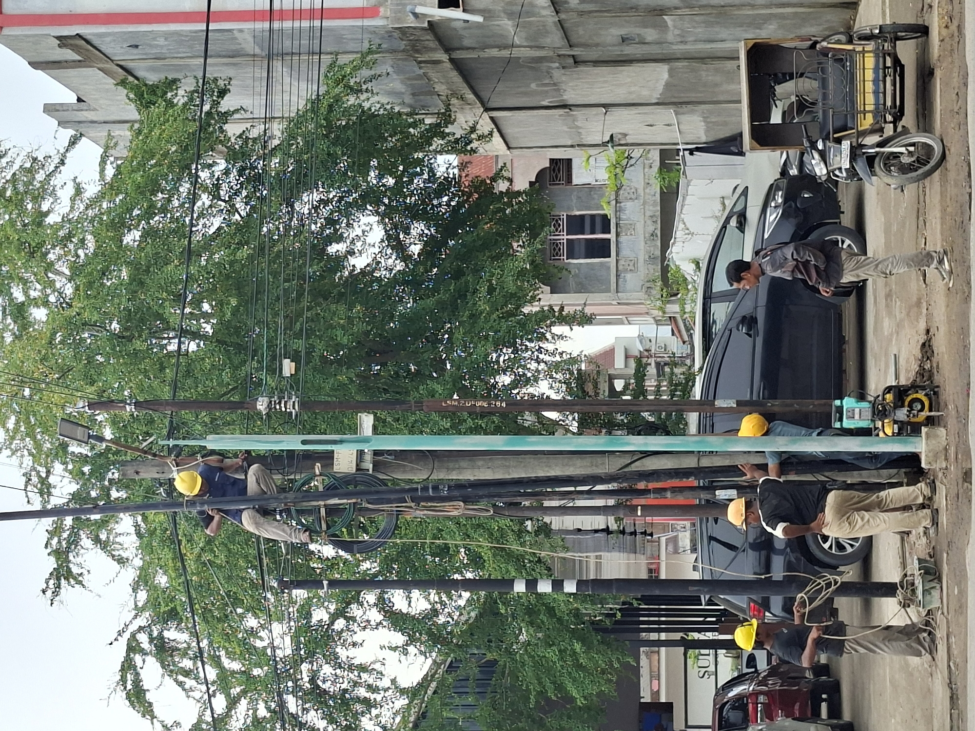 In Lhokseumawe, Indonesia, three men wearing yellow hard hats are working near utility poles on a street. One man is perched precariously on a pole, seemingly adjusting wires. Another man stands near the base of the pole, holding a coil of rope, while a third man stands beside him, his hand resting on a different pole. A portable generator sits on the ground near the poles, and a motor-rickshaw is parked to the right. Several vehicles, including a dark-colored SUV and a maroon car, are visible in the background, along with lush green trees and concrete buildings, suggesting an urban environment. The sky is overcast.