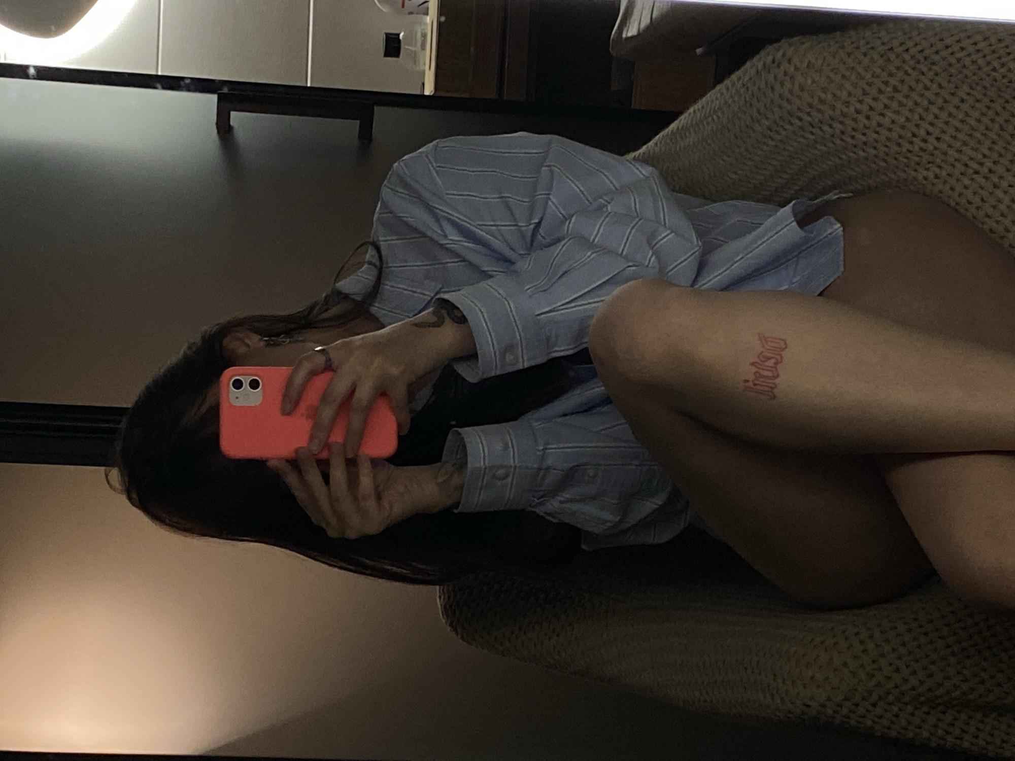 The image displays a mirror selfie of a person lying down in an indoor setting, rotated 90 degrees counter-clockwise from a natural upright orientation. The subject, with long dark hair, is positioned on a textured, light brown or beige surface, likely a blanket or upholstered furniture. They are holding a bright coral/orange smartphone with two visible camera lenses, with their hands and phone partially obscuring their face.
The person is wearing an oversized, light blue collared shirt with thin white stripes. On their left forearm, a dark, abstract tattoo, possibly resembling a snake or tribal pattern, is visible. A prominent red script tattoo, appearing to read "Jirúsa" in a gothic-style font, is located on their upper right thigh.
The room is dimly lit, suggesting an evening or nighttime scene, with a bright, circular artificial light source prominently reflected in the upper left corner of the mirror. The background reflected in the mirror is dark and indistinct, showing hints of furniture or room features. The overall atmosphere is casual and intimate. The location context is Votkinsk, Russia.