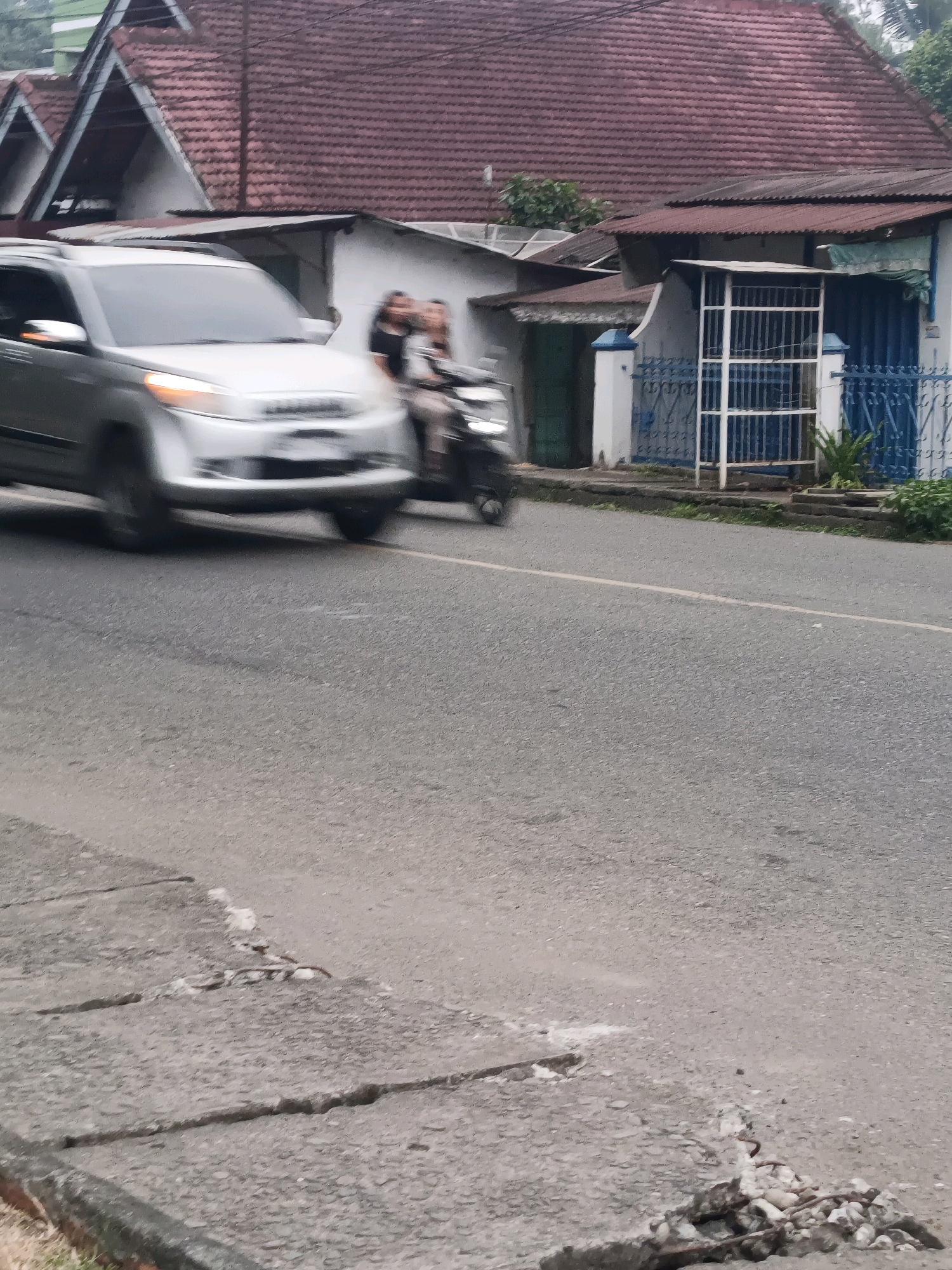 The image shows a street scene in Lubuklinggau, Indonesia. A silver SUV is moving down the road, its headlights on. A motorcycle is also visible, carrying two passengers. In the background, there are several buildings with red tile roofs, typical of Indonesian architecture. The street is paved with asphalt, and a white line marks the center. The environment appears to be an urban or suburban area, with some vegetation visible along the roadside.