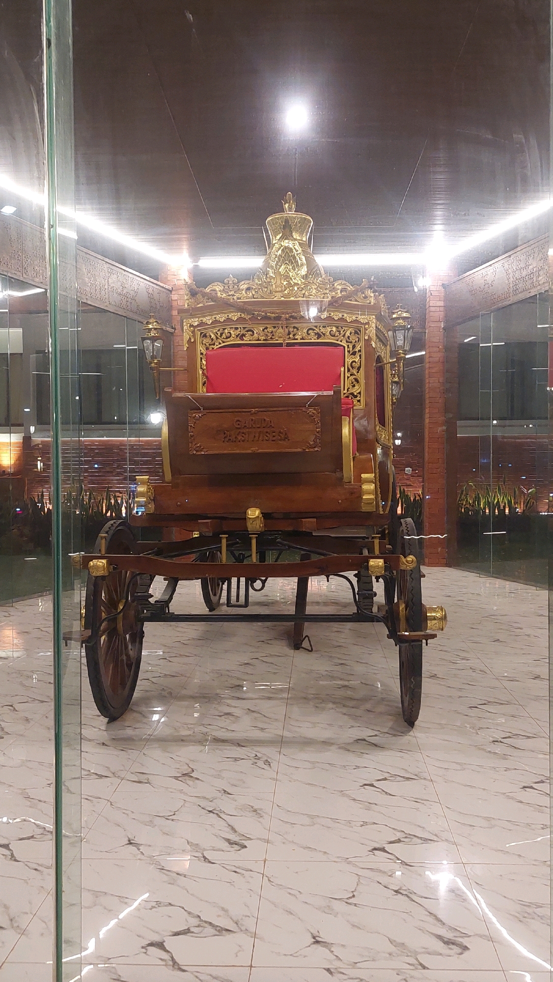 The image shows a photograph of an ornate wooden carriage, possibly a historical exhibit, situated indoors on a marble-tiled floor. The carriage is brown with gold and red accents and features decorative carvings. It has four wheels, two large wooden ones in the back and two smaller ones in the front. A sign on the side of the carriage reads "GARUDA PAKSI WISESA". The carriage is positioned centrally in the frame, with reflections visible on the glass walls surrounding it. The ceiling above is dark and appears to have strip lighting. The background suggests a modern building with brick walls and some greenery visible through the glass. This carriage is located at the Bogor Palace exhibition hall in Cibinong, Indonesia, a venue that showcases historical artifacts and royal heritage.