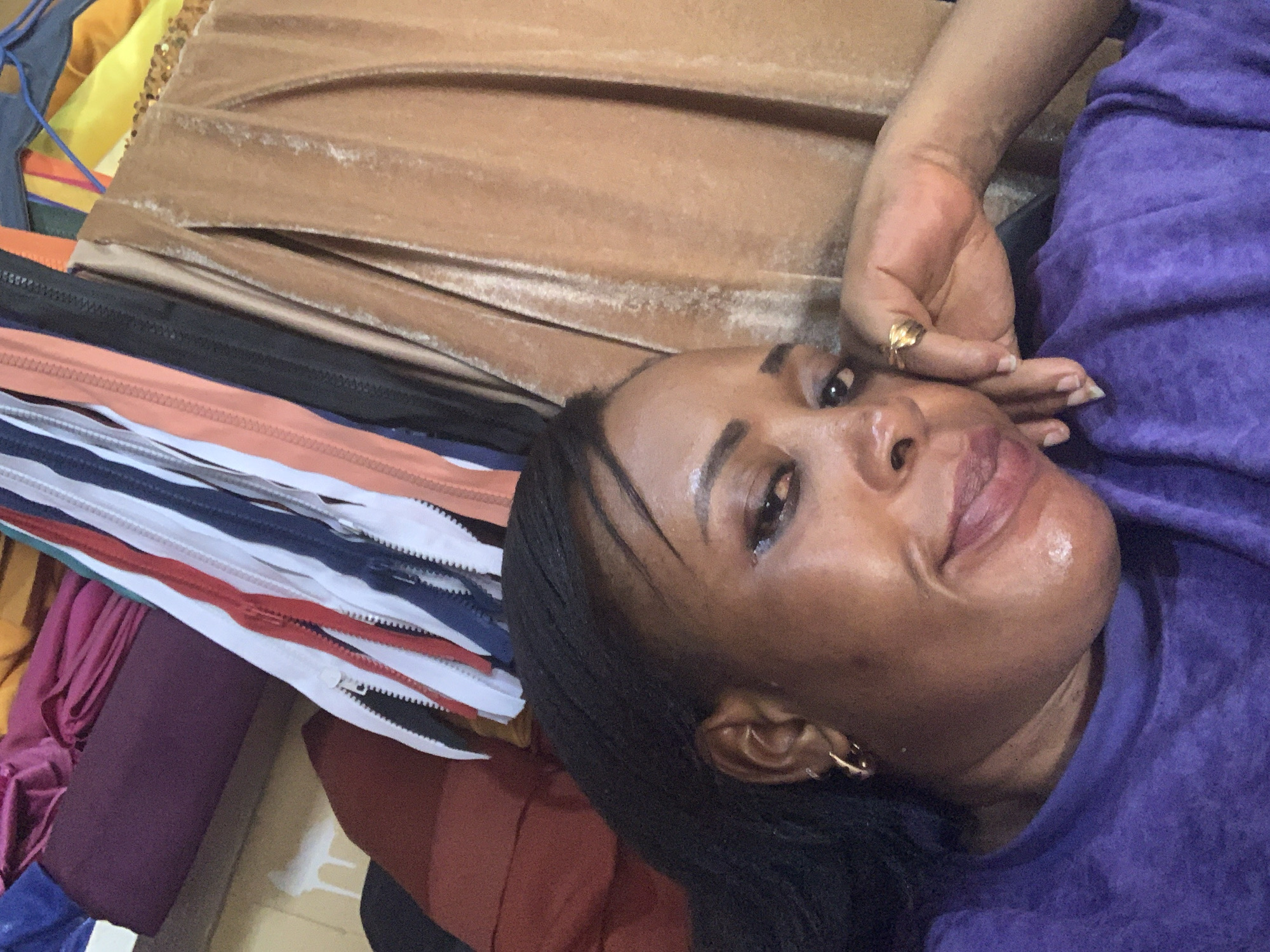 A close-up, overhead, selfie-style shot shows a Black woman resting her head on her hand. She is wearing a purple top and a gold hoop earring in her left ear. She has a gold ring on her right ring finger. Her hair is braided. The background consists of fabric and zippers. The scene is likely indoors in Sabon Tasha, Nigeria.