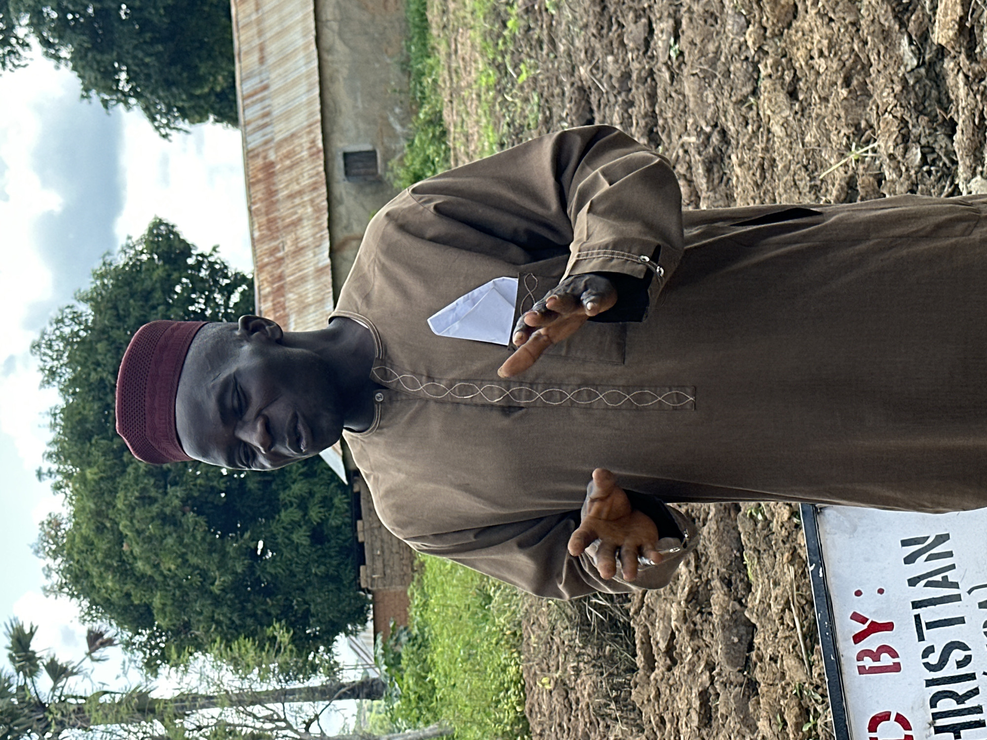 In the city of Afogo, Nigeria, a man stands in a field. He is wearing a brown robe and a red cap. His mouth is open as if he is speaking, and his hands are raised. There is a white cloth in his breast pocket. In the background, there is a building with a corrugated metal roof and trees. A sign in the foreground reads "BY: CHRISTIAN". The ground is covered in dirt and some sparse vegetation. The sky is cloudy.