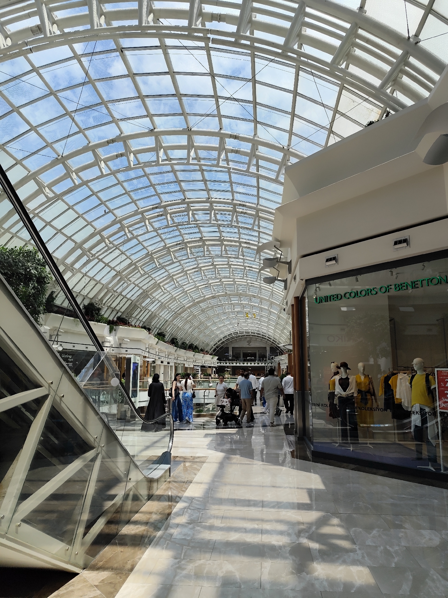 The image shows the interior of a shopping mall in Sarıyer, Turkey. The location is identifiable by the "UNITED COLORS OF BENETTON" storefront signage, as well as storefronts in the distance of the image. The mall features a large, arched glass roof supported by a white framework, allowing natural light to flood the interior. People are walking through the mall, including individuals, couples, and families, and a woman with a stroller. An escalator is visible on the left side of the image. The floor is marble-like. Storefronts line the mall, and mannequins are visible in the Benetton store window.
