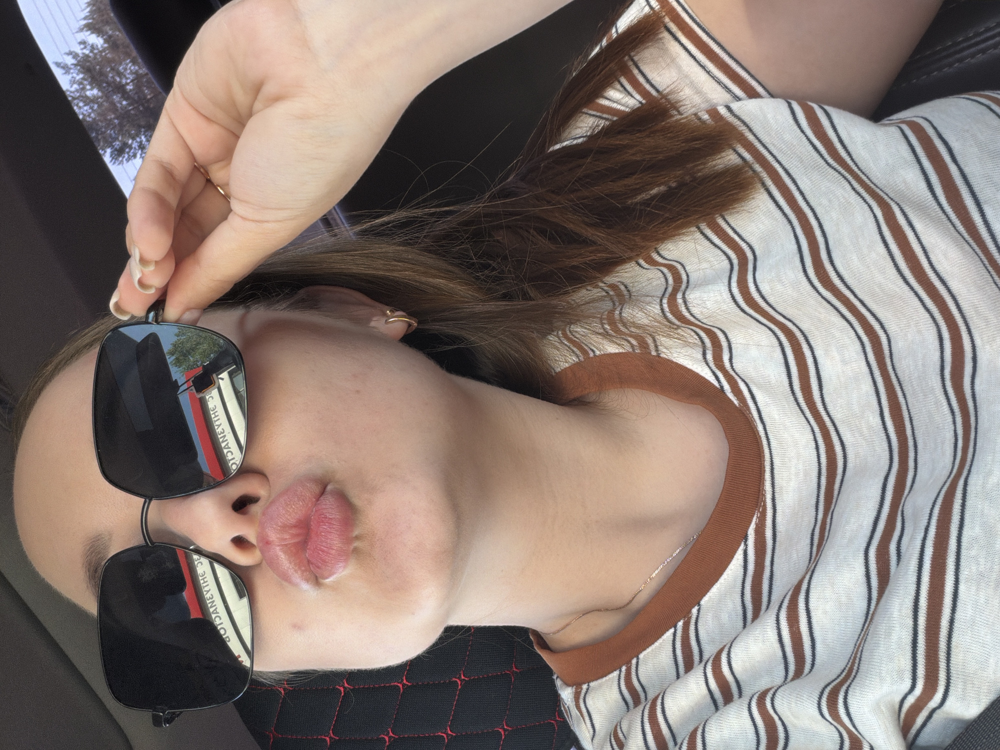A person, possibly female based on facial features and a visible gold hoop earring on the left ear, is photographed from a close-up perspective. The person is wearing sunglasses with dark lenses that reflect a cityscape, potentially within the city of Chaikovsky, Russia. The reflection shows buildings with signage written in what appears to be Russian. The person is puckering their lips. They are wearing a white t-shirt with brown and dark stripes and a brown collar. Part of the person's hair is visible, appearing auburn. The person's left hand holds the sunglasses. The background includes a dark-colored car interior, including a seat cover with a red diamond pattern. Outside the car, some trees can be seen.