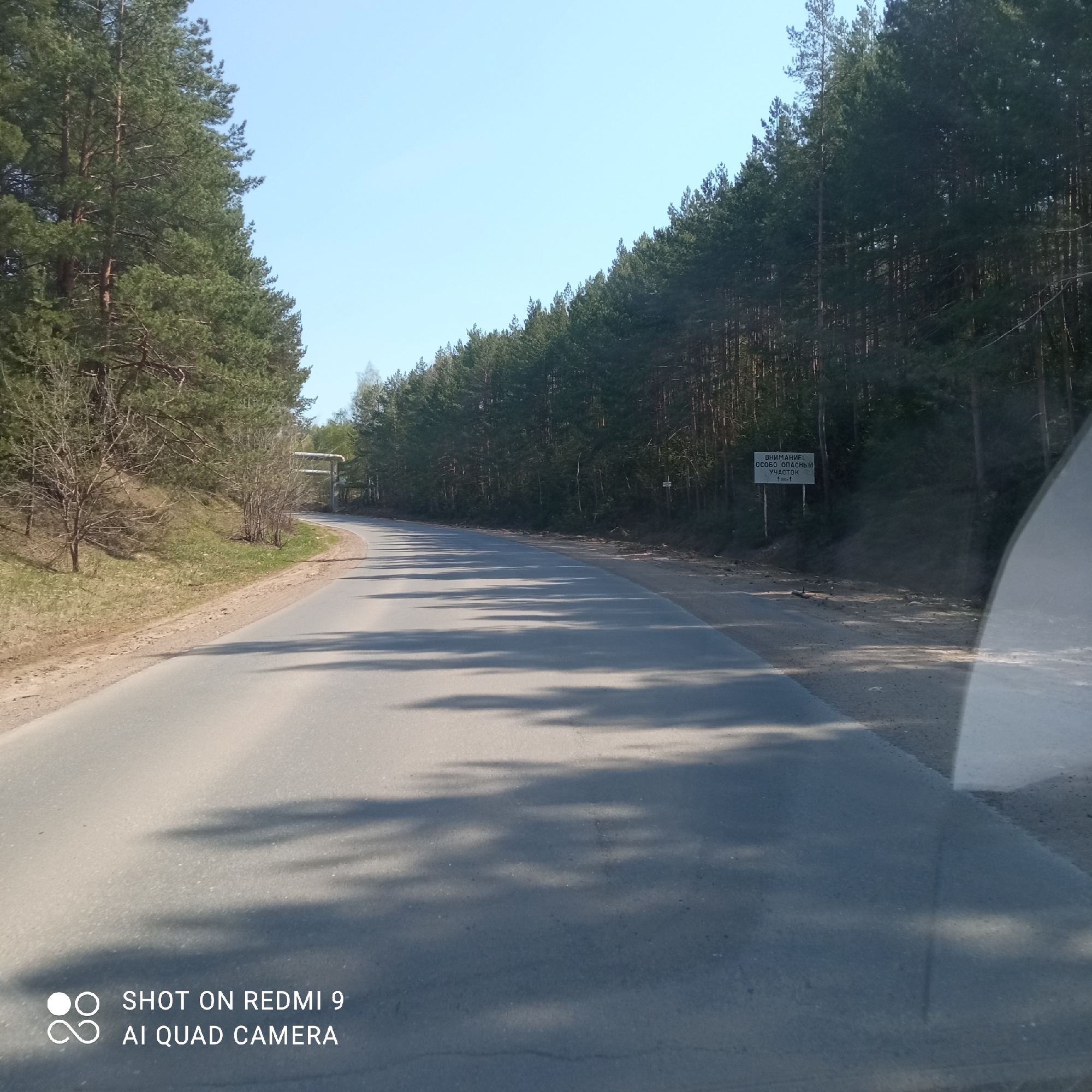 A road stretches through a forest area. The road is paved with asphalt, and shadows from the trees above fall across its surface. Trees line both sides of the road. A sign in Russian is visible on the right side of the road. In the distance, a metal structure spans the road. The image includes the text "SHOT ON REDMI 9 AI QUAD CAMERA." The location is Прикамский, Russia.