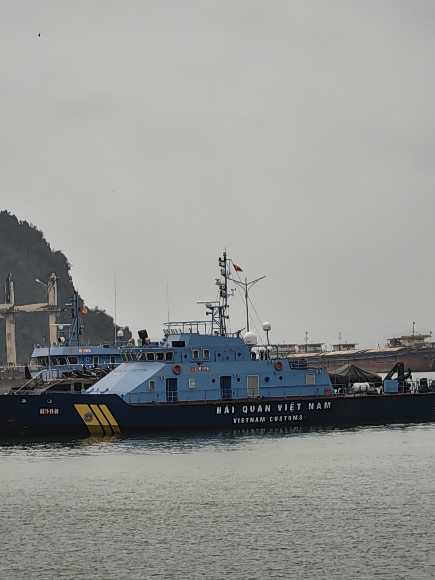 The image shows a blue Vietnam Customs boat, labeled "HQ 11-01-06" on the bow and "HẢI QUAN VIỆT NAM VIETNAM CUSTOMS" on the hull, docked in the waters off Hong Gai Ward, Vietnam. Another similar boat is partially visible behind it, along with other larger vessels and a coastal mountain. The sky is overcast.