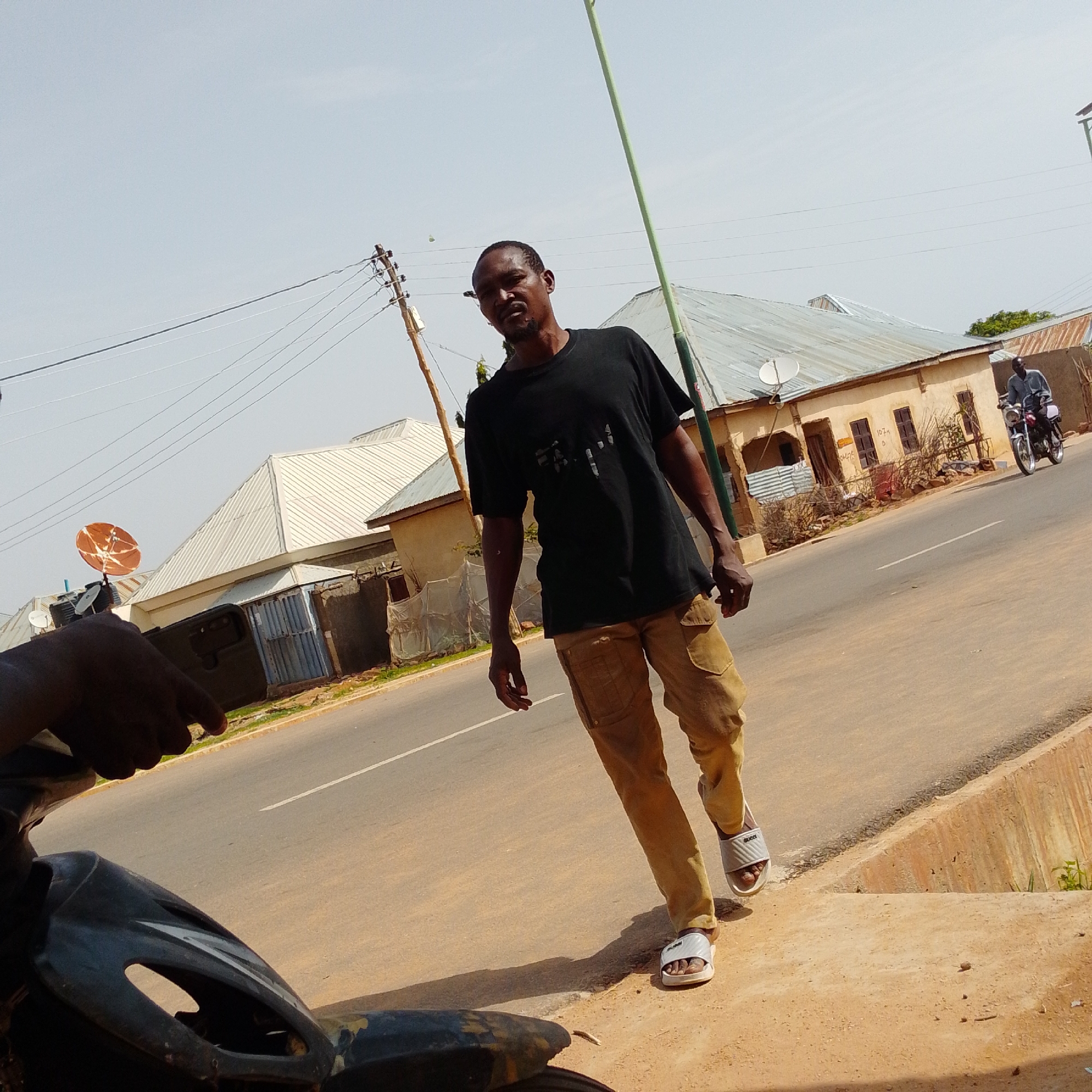 A man in a black t-shirt and tan cargo pants walks along a paved road in Gombe, Nigeria. He is wearing white slide sandals. A motorcycle with a rider is visible in the background, traveling in the same direction. The road is lined with buildings that have corrugated metal roofs. A satellite dish is mounted on one of the buildings. Power lines are strung across the sky, supported by utility poles. The sky is clear and blue, suggesting it is daytime. The man appears to be walking casually. A portion of a dark-colored motorcycle is visible in the foreground, from the perspective of someone sitting on it. The overall scene depicts a typical street in a Nigerian town.