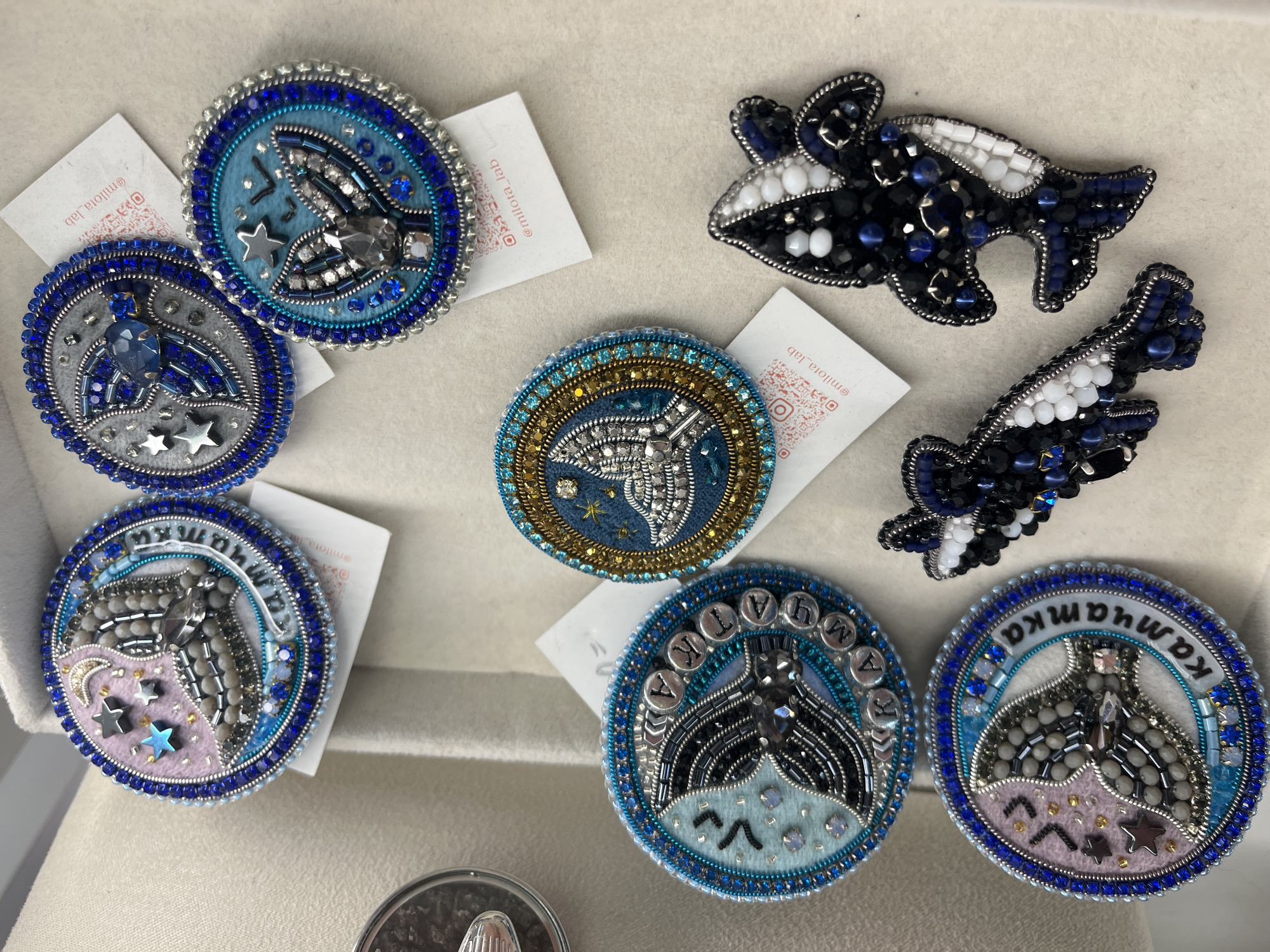 The image shows a collection of beaded brooches arranged on a beige surface, possibly a display for crafts. The brooches are circular or shaped like marine animals, featuring intricate beadwork in shades of blue, silver, gold, pink, and white. Many of the brooches depict whale tails and stars. Some circular brooches have text embroidered around the edges in Cyrillic letters. Attached to some of the brooches are small white tags with QR codes and social media handles, suggesting they are for sale or display by a craftsperson. There are no people or animals visible in the image, nor is there any indication of the location being in the city of Svetly, Russia. The environment is a close-up shot, focusing solely on the brooches.
