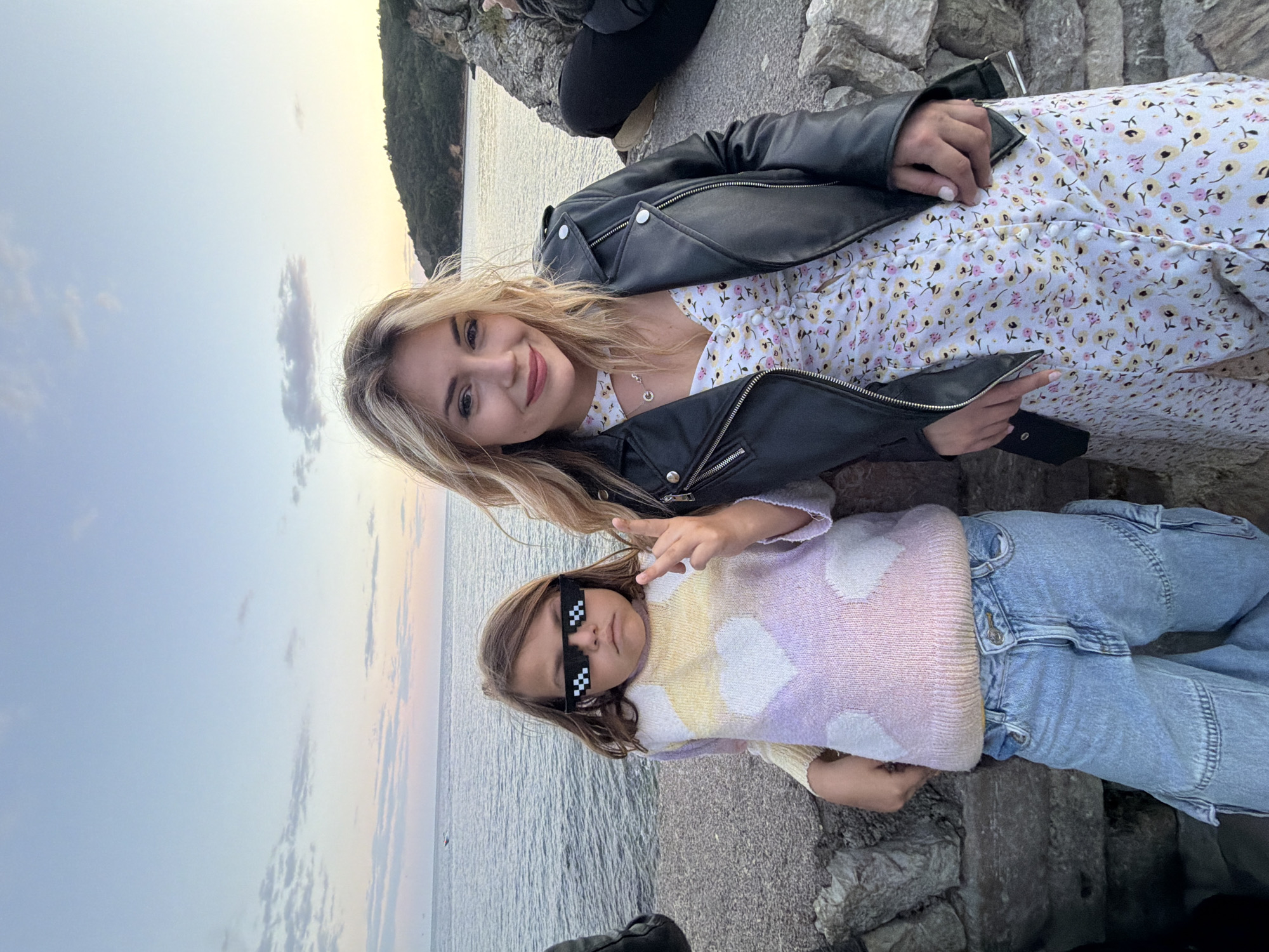 The image shows two individuals, an adult and a child, outdoors at the location of Budva, Montenegro. The adult, a blonde-haired woman, is wearing a floral dress with a black leather jacket. The child has brown hair and is wearing a light-colored sweater with cloud-like patterns and light blue jeans and pixelated sunglasses. They are positioned near a body of water with a rocky landscape in the background. The sky appears to be at dusk or dawn, with soft colors. They are posing on a low stone wall. The child is gesturing with their hand.