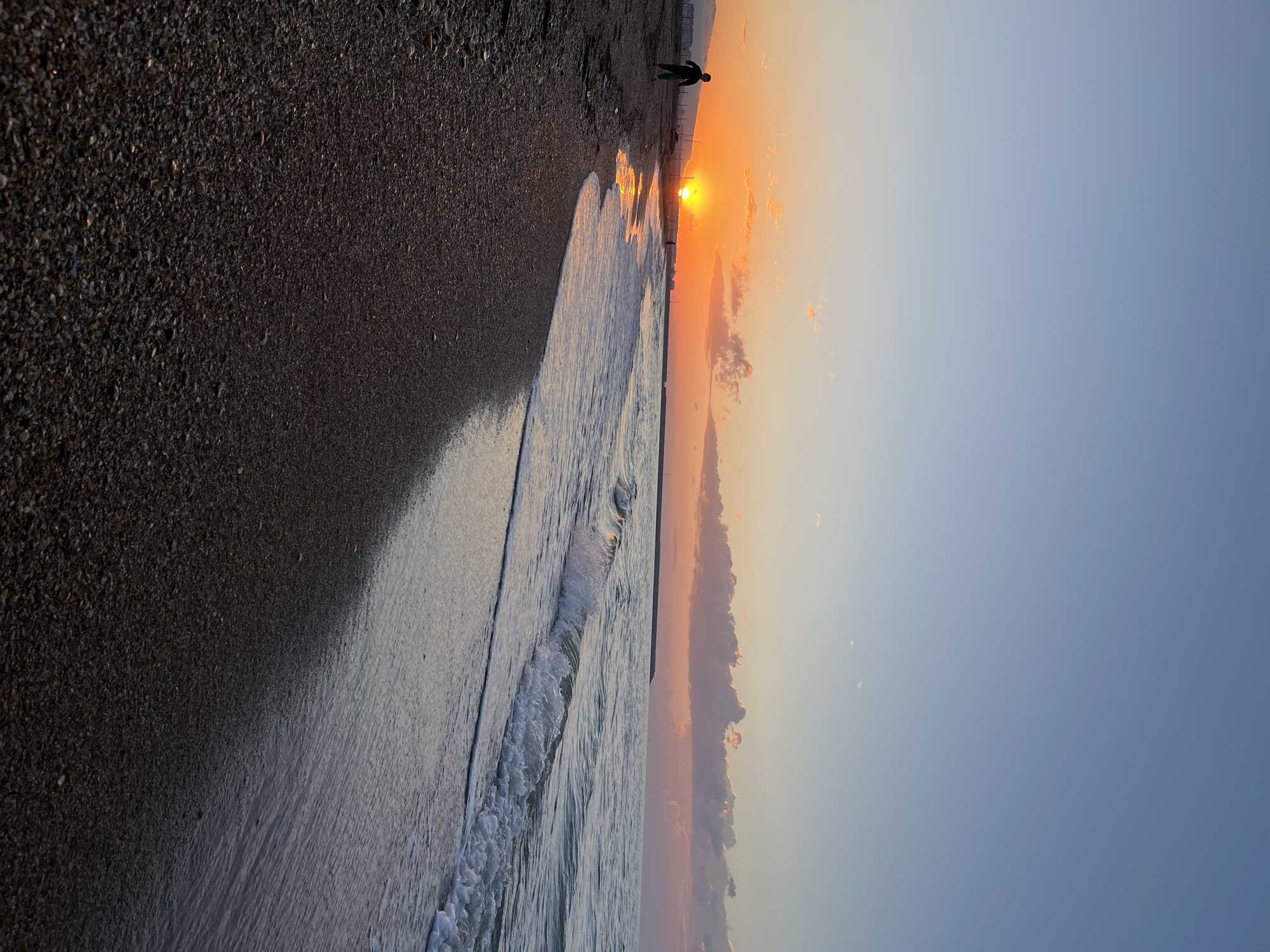 The image depicts a beach scene at sunset in Kaspiysk, Russia. The beach is dark and coarse. Waves from the Caspian Sea are breaking on the shore. A solitary figure stands on the beach in the distance, silhouetted against the setting sun. The sun is positioned low on the horizon, casting a warm orange glow across the sky, which transitions to a light blue. Some clouds are present in the orange-hued area of the sky.