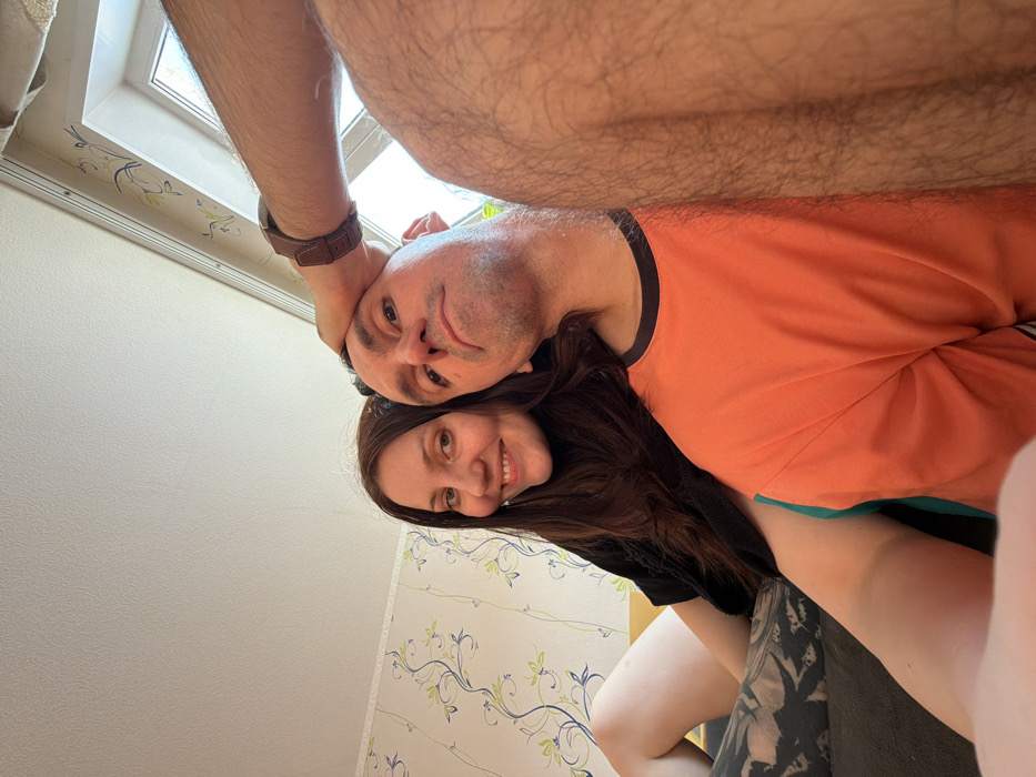 This image depicts an indoor residential scene featuring two individuals, with a third person partially visible. The location, stated as Chaikovsky, Russia, cannot be independently verified from the visual information within the image.
**People:**
1. **Female:** Positioned on the left, a female with long, dark brown hair is smiling and looking towards the camera. She is wearing a black top. Her left leg, with fair skin, is partially visible and features a large, dark, intricate tattoo on the upper thigh/hip area.
2. **Male (center):** Positioned centrally, a male with a largely bald head and short dark hair on the sides is looking directly at the camera with a neutral expression. He is wearing an orange t-shirt with a black collar and a vertical teal stripe along the side.
3. **Third Person (partially visible):** On the far right, the right arm and right leg of a third individual are visible. The arm is raised, with a brown leather-strap watch on the wrist, and the hand is resting on the top of the central male's head. The right leg is heavily covered in dark hair.
**Actions and Interactions:** All visible individuals are positioned closely, suggesting an intimate or casual setting. The individuals are looking towards the camera, indicative of a selfie-style photograph. The third person's hand resting on the central male's head implies physical interaction.
**Environment and Objects:**
* **Walls:** The background wall is covered with light-colored wallpaper featuring a repeating pattern of green and blue floral or vine motifs.
* **Furniture:** A yellow surface, possibly the back of a sofa, is visible behind the female. Dark grey or black fabric, likely a sofa cushion, is visible in the foreground.
* **Window:** A white-framed window, allowing natural light into the room, is present in the upper right background.
* **Ceiling:** A plain white ceiling is visible at the top of the frame.
* **Clothing/Accessories:** The central male wears an orange t-shirt. The female wears a black top. The partially visible third person wears a brown leather-strap watch.