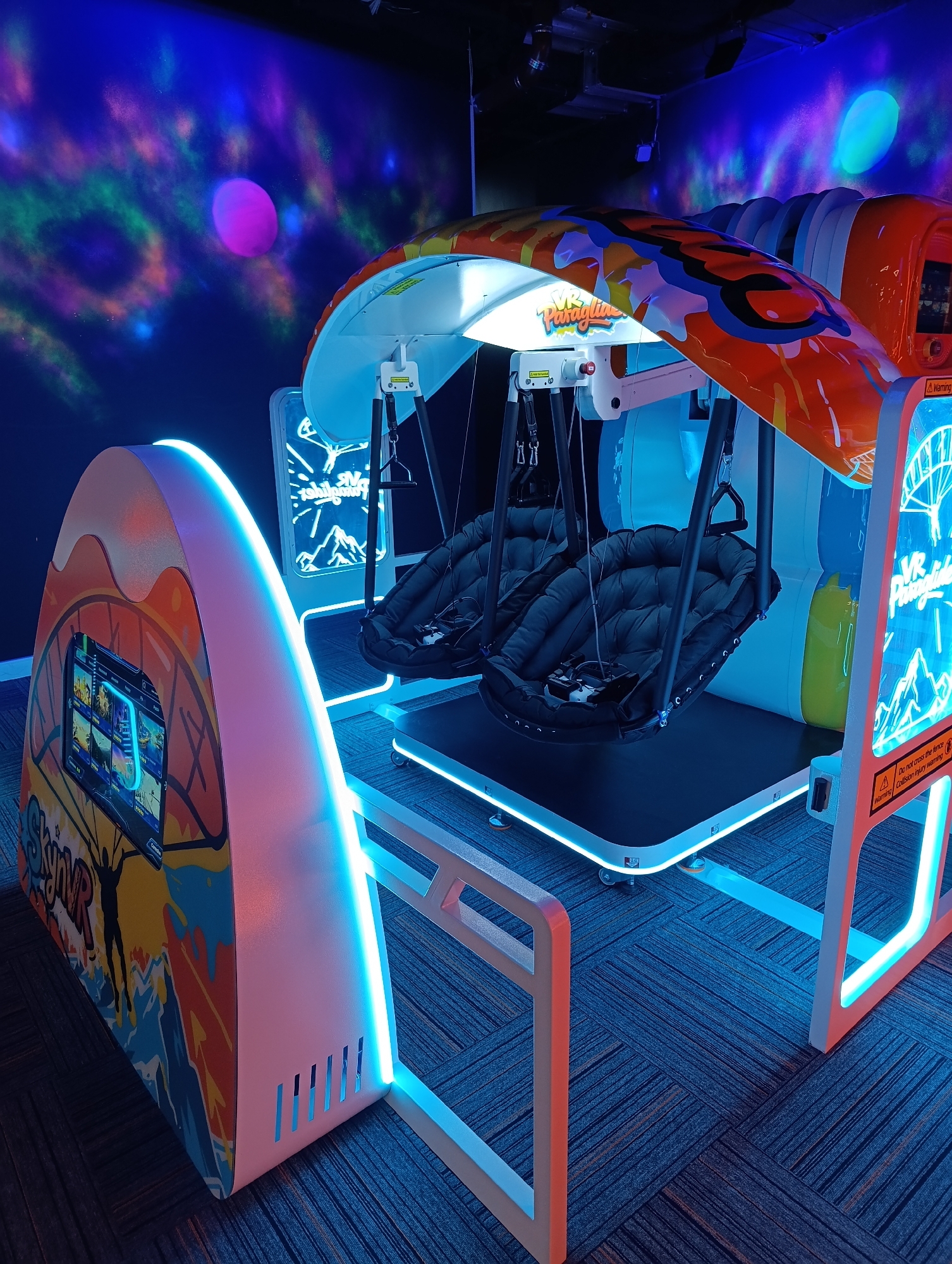 In a room with a dark blue carpet, a VR paragliding simulation machine is set up. The machine features two black cushioned seats suspended from an overhead structure. The overhead structure is adorned with a vibrant orange, blue, and yellow graphic design. To the left of the main unit is a smaller, curved console with a touchscreen display showing various images, and is also decorated with similar graphics. The walls behind the machine are painted dark blue and illuminated with projected nebulae-like patterns in purple, green, and pink, creating a cosmic atmosphere. The scene is further enhanced by blue LED strip lighting around the base of the VR machine and the consoles. The overall environment suggests an entertainment or arcade setting, specifically a VR experience center, though no specific location in Eyüpsultan, Turkey is identifiable from the image. There are no people present in the image.