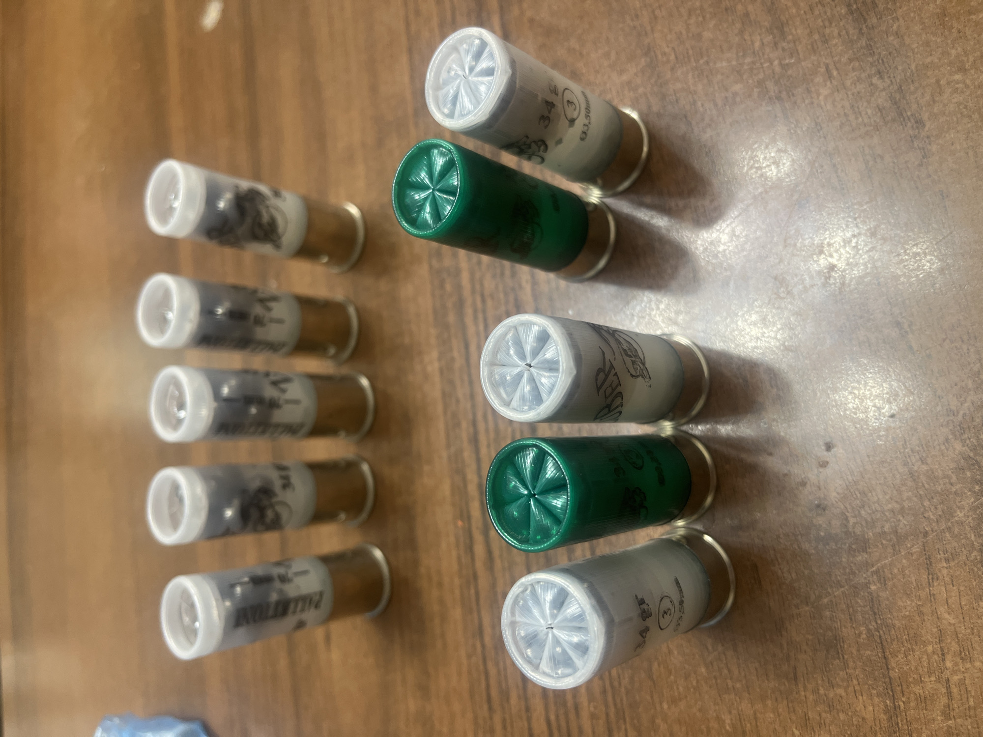 Ten shotgun shells are neatly arranged on a wooden surface, suggesting an indoor setting. Six of the shells are predominantly white or light gray, while the other four are dark green. All shells feature a clear plastic end with a distinct star-crimp pattern and a metallic, brass-colored base.
The shells are organized into two vertical columns. The left column contains six white/gray shells, stacked with their clear ends facing upwards. The right column holds four shells, alternating in color: a white/gray shell at the top, followed by a green, then another white/gray, and finally a green shell at the bottom, also with their clear ends visible.
Visible text on the white/gray shells includes "34g", a circled "3", and "93,50mm". Partially visible text also shows "PALLENTONI" (Italian for buckshot) and "SER". Text on the green shells is less clear but also appears to indicate weight or shot size. The wooden surface has a visible grain and some minor scuffs.